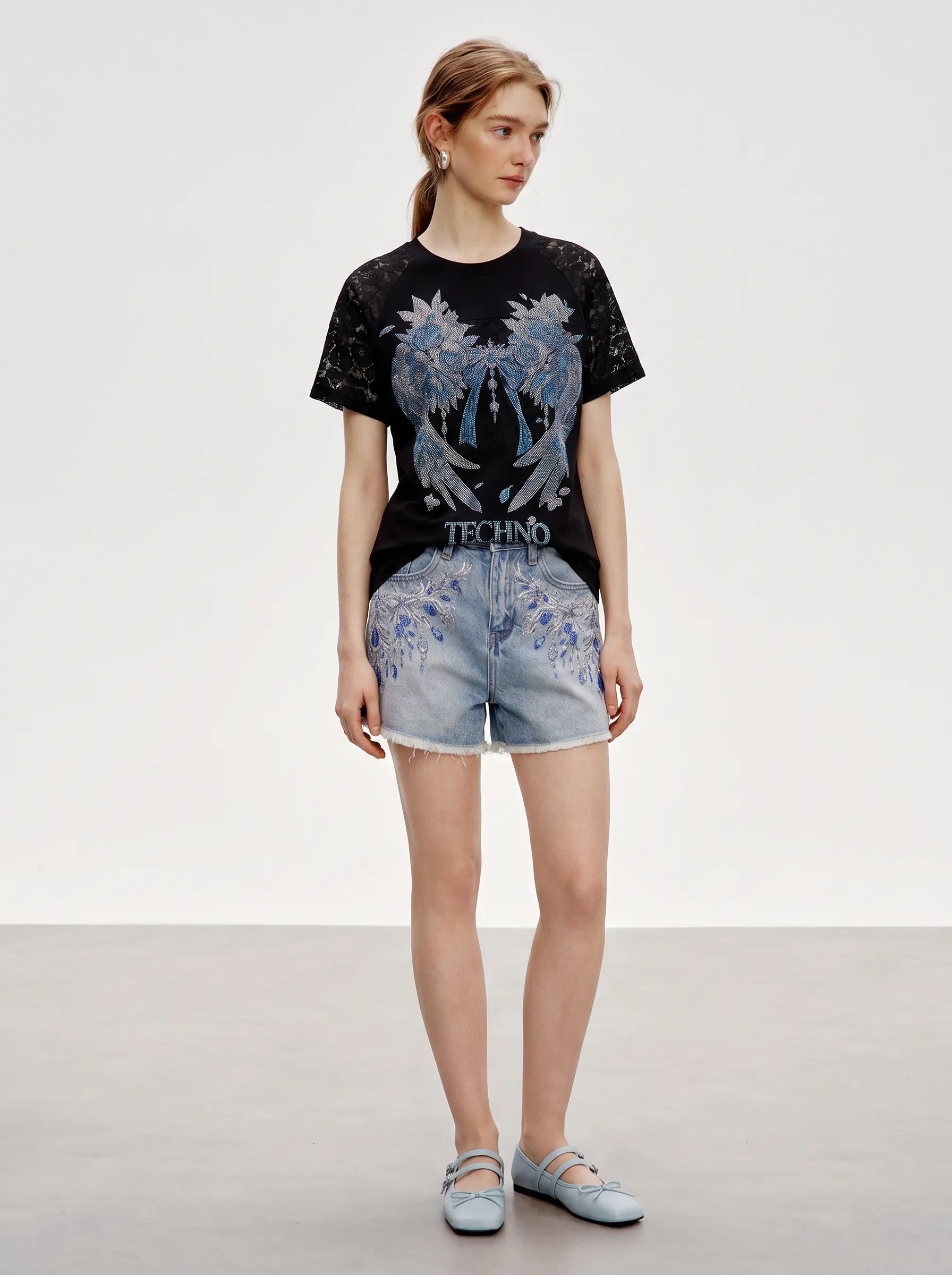 A model stands in a studio wearing a black short-sleeve t-shirt with a large blue angel graphic and the word 'TECHINO' on the front, paired with light blue denim shorts and light blue flats. TCH Rhinestone Floral Wing Techno Graphic T-Shirt