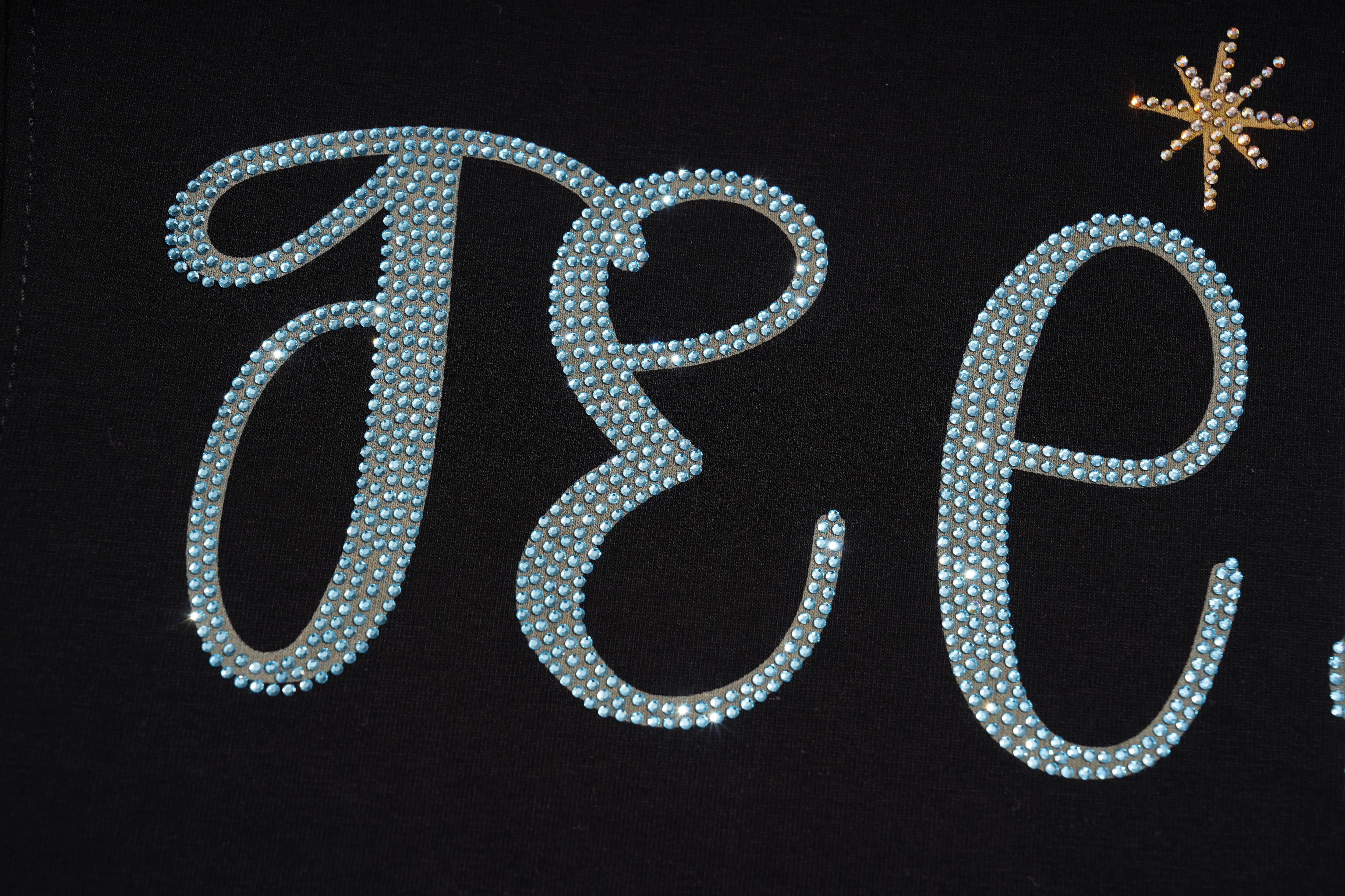 A close-up of a black fabric surface featuring large cursive letters outlined with light blue rhinestones, with a small gold star embellishment in the upper right. TCH Rhinestone Galaxy Spiral Graphic Black T-Shirt