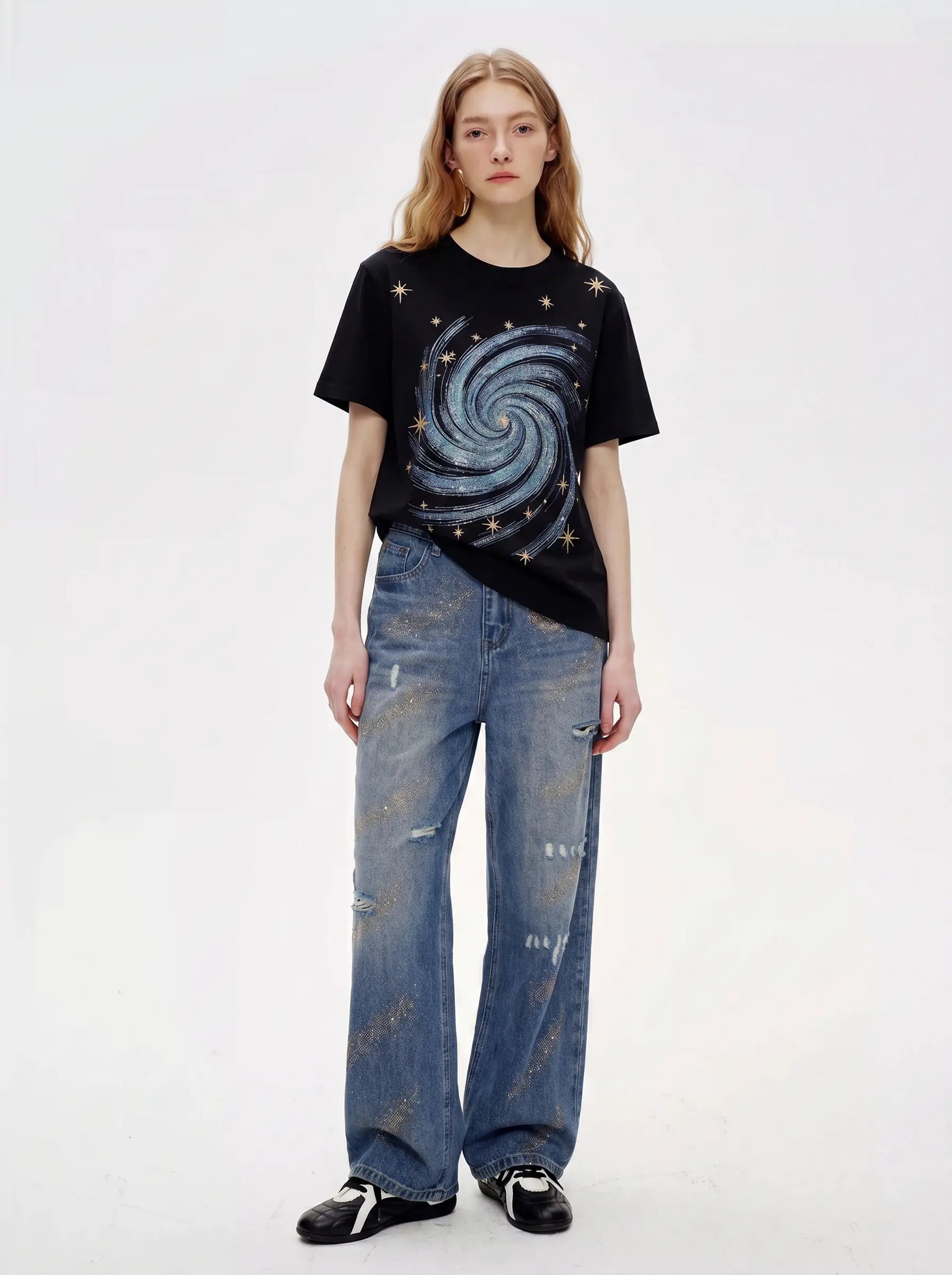 A model stands against a plain white studio background wearing a black graphic T-shirt with a blue spiral galaxy print and small gold stars, paired with distressed blue jeans and black-and-white sneakers. TCH Rhinestone Galaxy Spiral Graphic Black T-Shirt