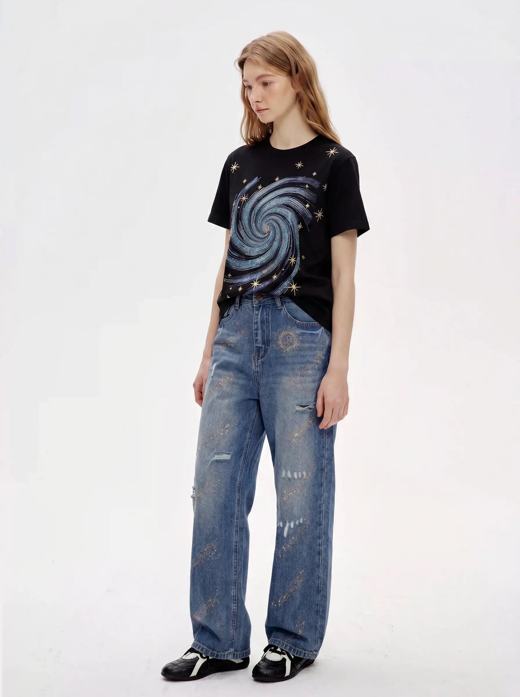 A model wearing a black short-sleeve t-shirt with a blue and silver galaxy graphic and distressed blue jeans, standing against a plain white studio background. TCH Rhinestone Galaxy Spiral Graphic Black T-Shirt