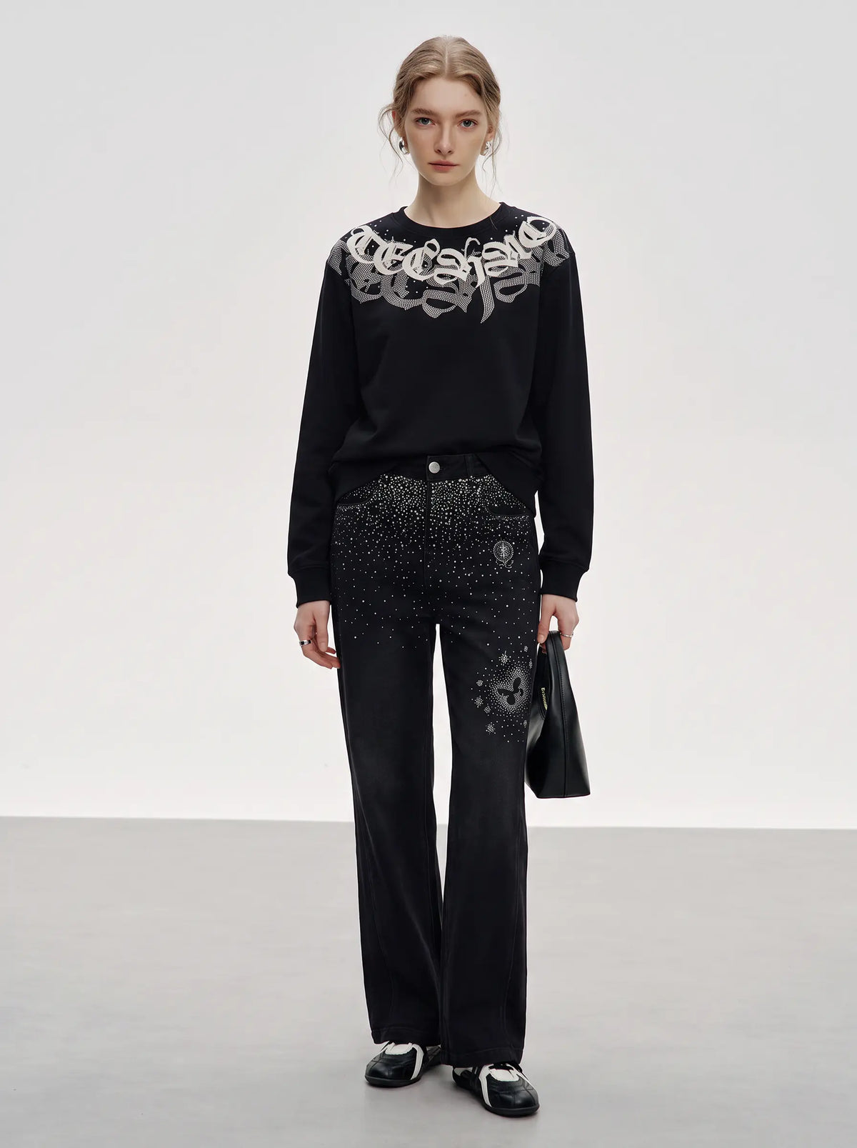 A model stands in a studio wearing a black long-sleeve top with a large silver graphic across the shoulders and black wide-leg pants covered in silver studs and embellishments. She is holding a black handbag against a clean light gray background. TCH Rhinestone Graphic Black Crewneck Sweatshirt