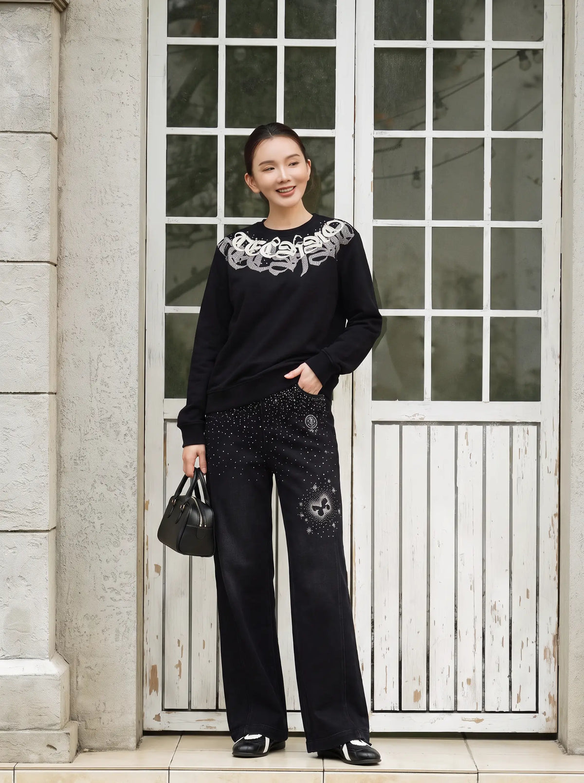 A woman stands in front of a white paneled door wearing a black long-sleeve top with a white graphic print across the chest and black wide-leg pants with small white speckles and skull details. She is holding a small black handbag. TCH Rhinestone Graphic Black Crewneck Sweatshirt