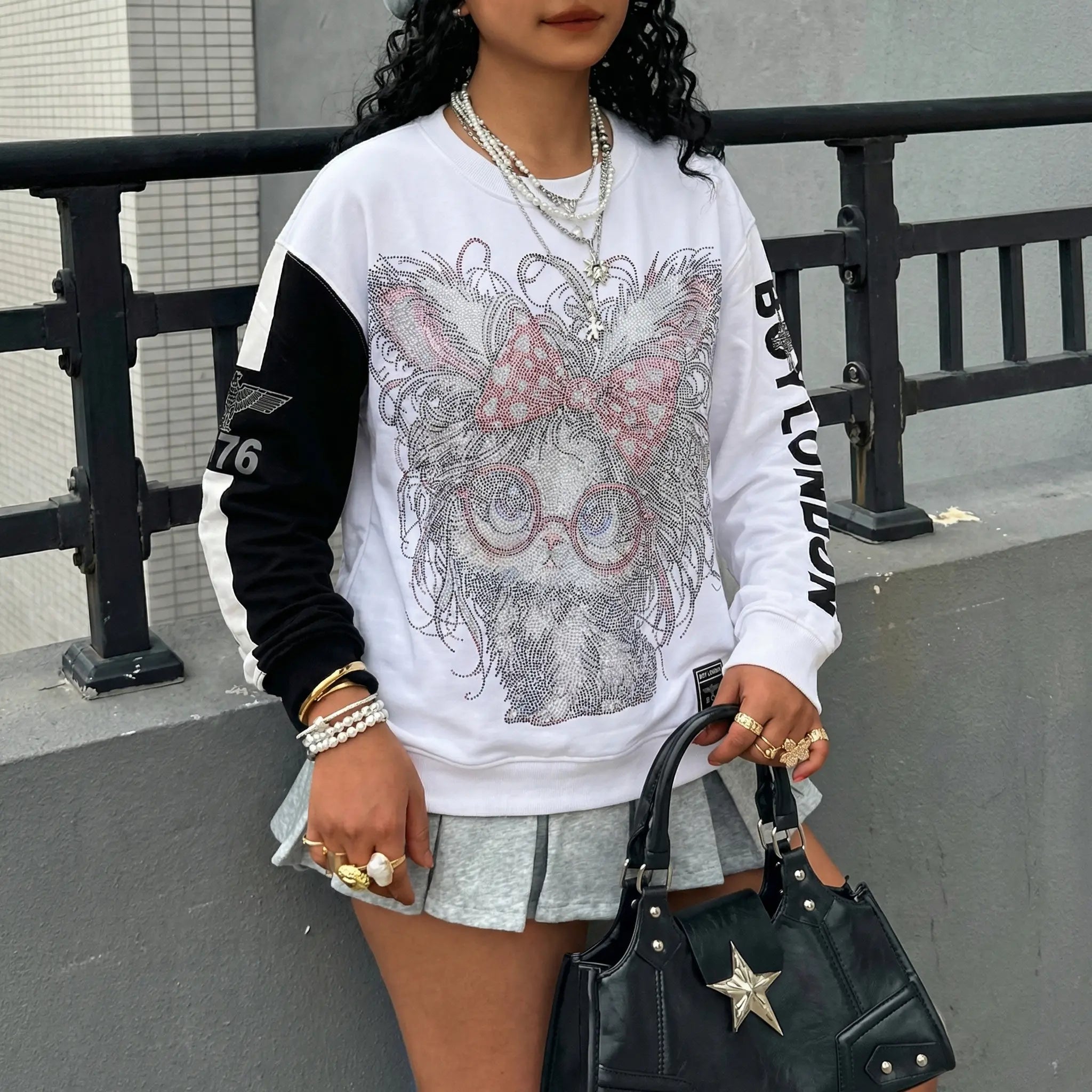 Person wearing a white sweatshirt with a graphic design, holding a black handbag. TCH Rhinestone Hello Kitty BOYLONDON Sweatshirt