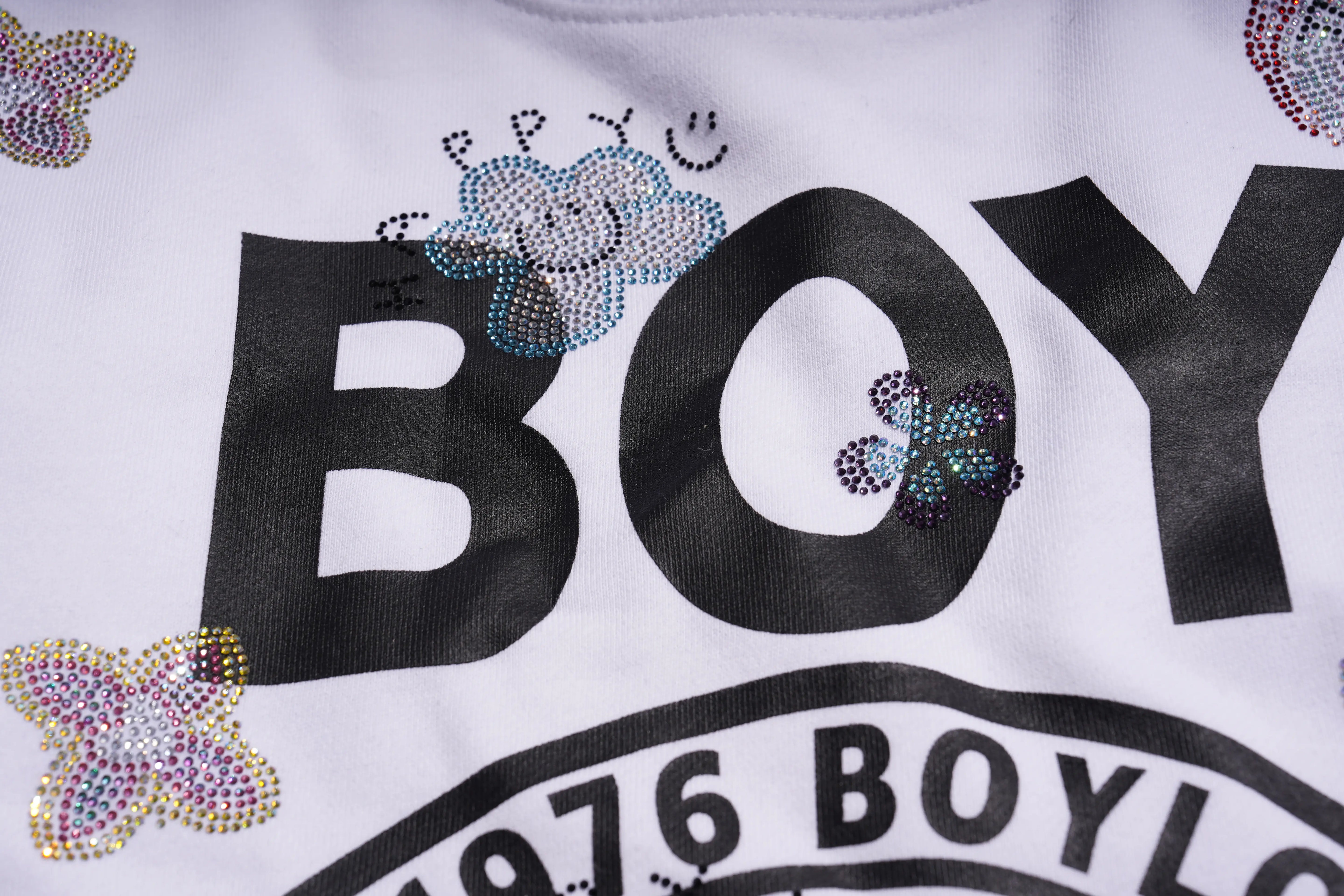 Close-up of a white shirt with 'BOY' text and decorative elements. TCH Rhinestone Logo Print Crew Neck Sweatshirt