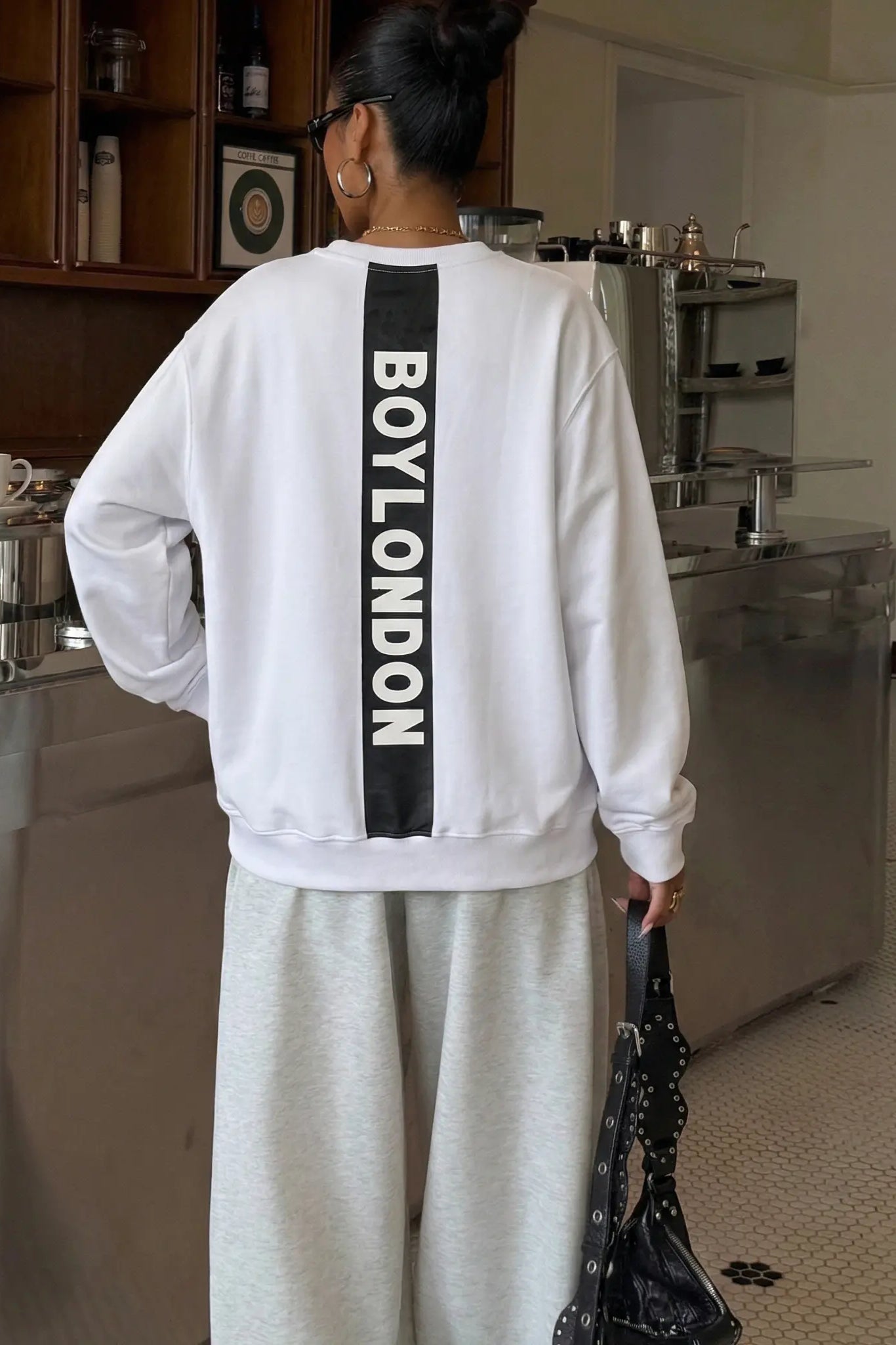 Person wearing a white sweatshirt with 'BOYLONDON' on the back in a kitchen setting. TCH Rhinestone Logo Print Crew Neck Sweatshirt