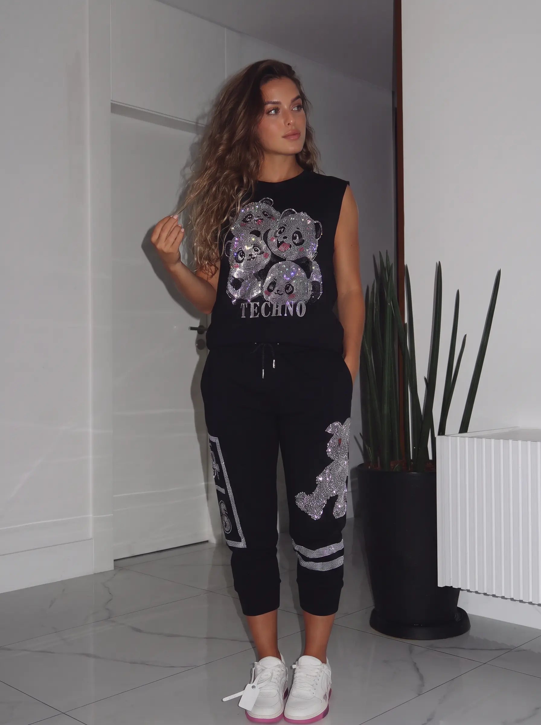 A woman modeling a black sleeveless Techno sweatshirt and matching jogger pants with silver sequined teddy bear graphics, standing in a modern indoor setting with white walls and a potted plant. TCH Rhinestone Panda Graphic Sleeveless T-Shirt
