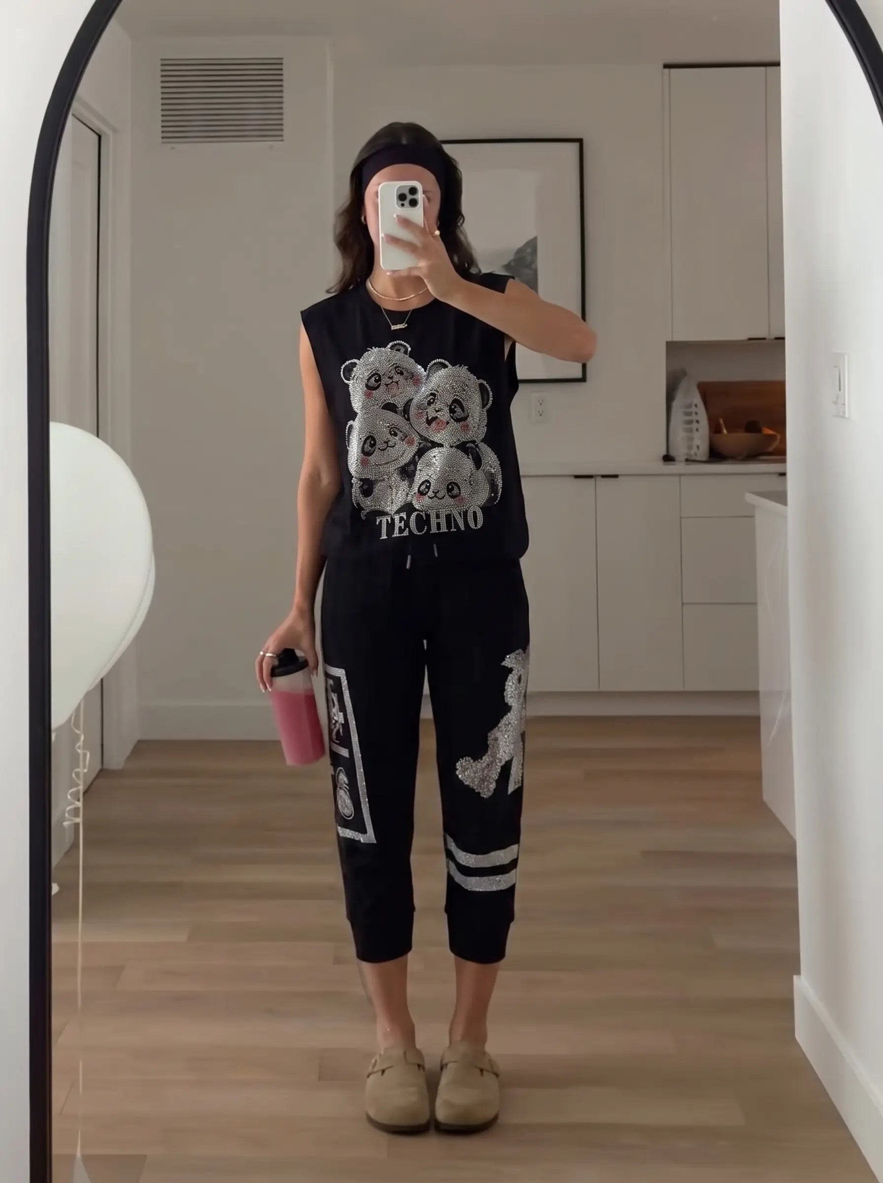 A person takes a mirror selfie in a home setting, wearing a black sleeveless top with a white panda graphic and the word 'TECHNO' on the front, paired with black jogger pants featuring white graphic details. They are wearing beige slip-on shoes and holding a pink cup. TCH Rhinestone Panda Graphic Sleeveless T-Shirt