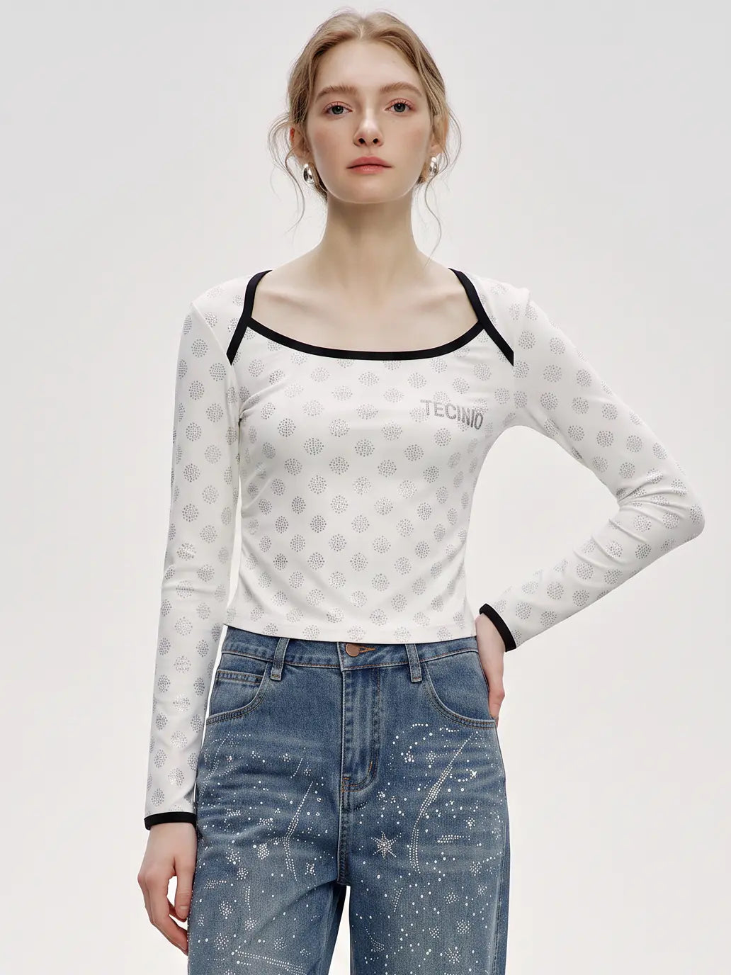 A model wearing a white long-sleeve top with a gray geometric pattern, black trim, and a square neckline, paired with blue jeans, against a light gray studio background. TCH Rhinestone Pattern Long Sleeve Top