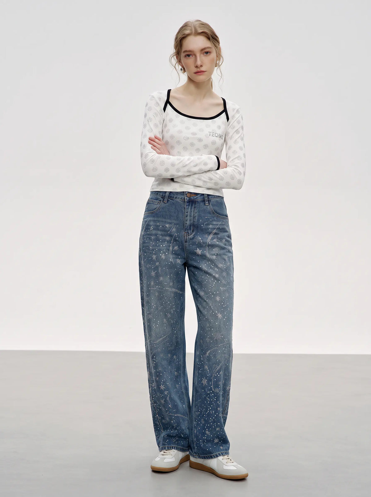A model stands in a studio wearing a white long-sleeve top with black trim and small dark motifs, paired with wide-leg blue jeans featuring a white abstract print, and white sneakers. TCH Rhinestone Pattern Long Sleeve Top