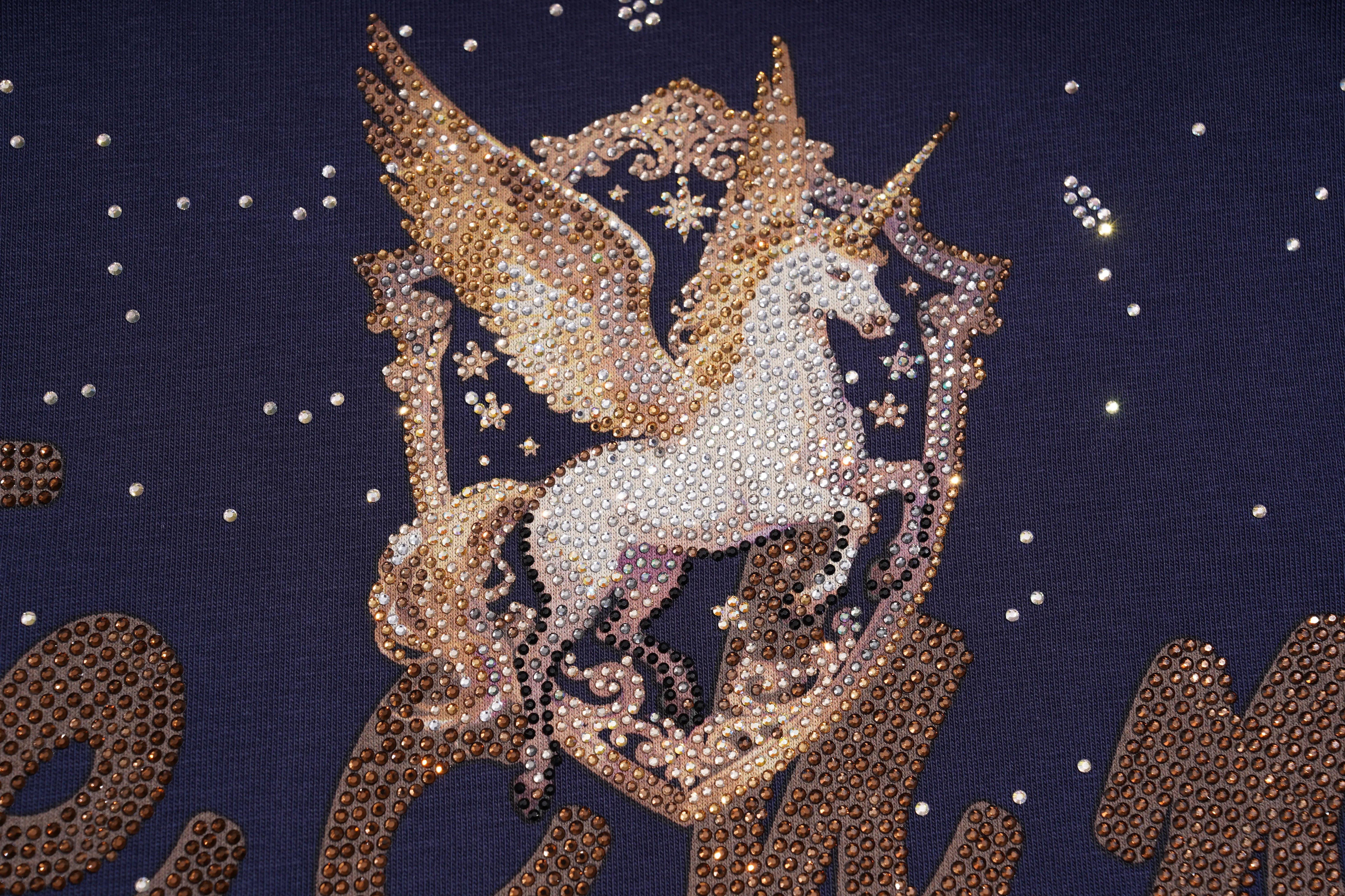 A close-up of a dark navy fabric featuring a large, detailed rhinestone design of a white and gold Pegasus unicorn within a shield, surrounded by small sparkling rhinestones. TCH Rhinestone Pegasus Wing Graphic Navy Blue T-Shirt