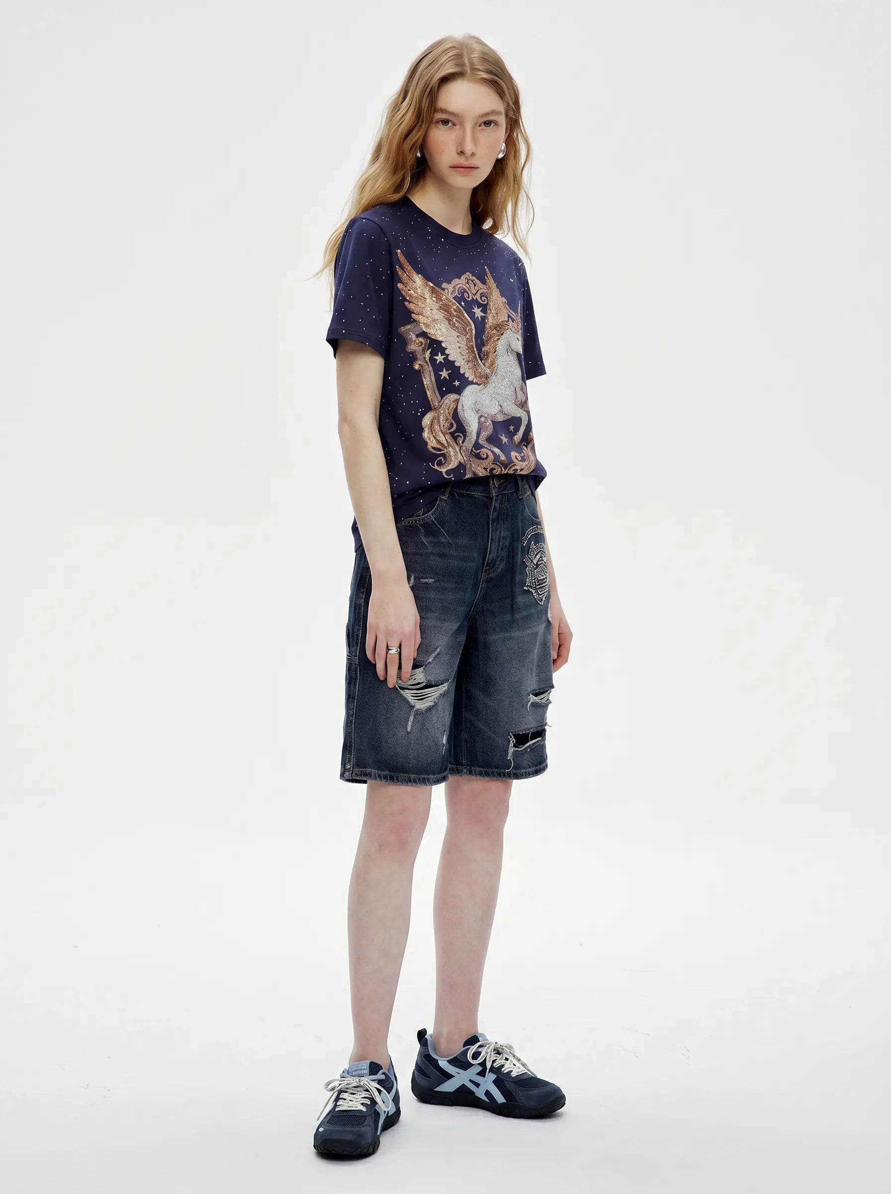 A model stands against a plain light gray studio background wearing a dark navy graphic T-shirt with a large eagle print, distressed denim shorts, and dark sneakers. TCH Rhinestone Pegasus Wing Graphic Navy Blue T-Shirt
