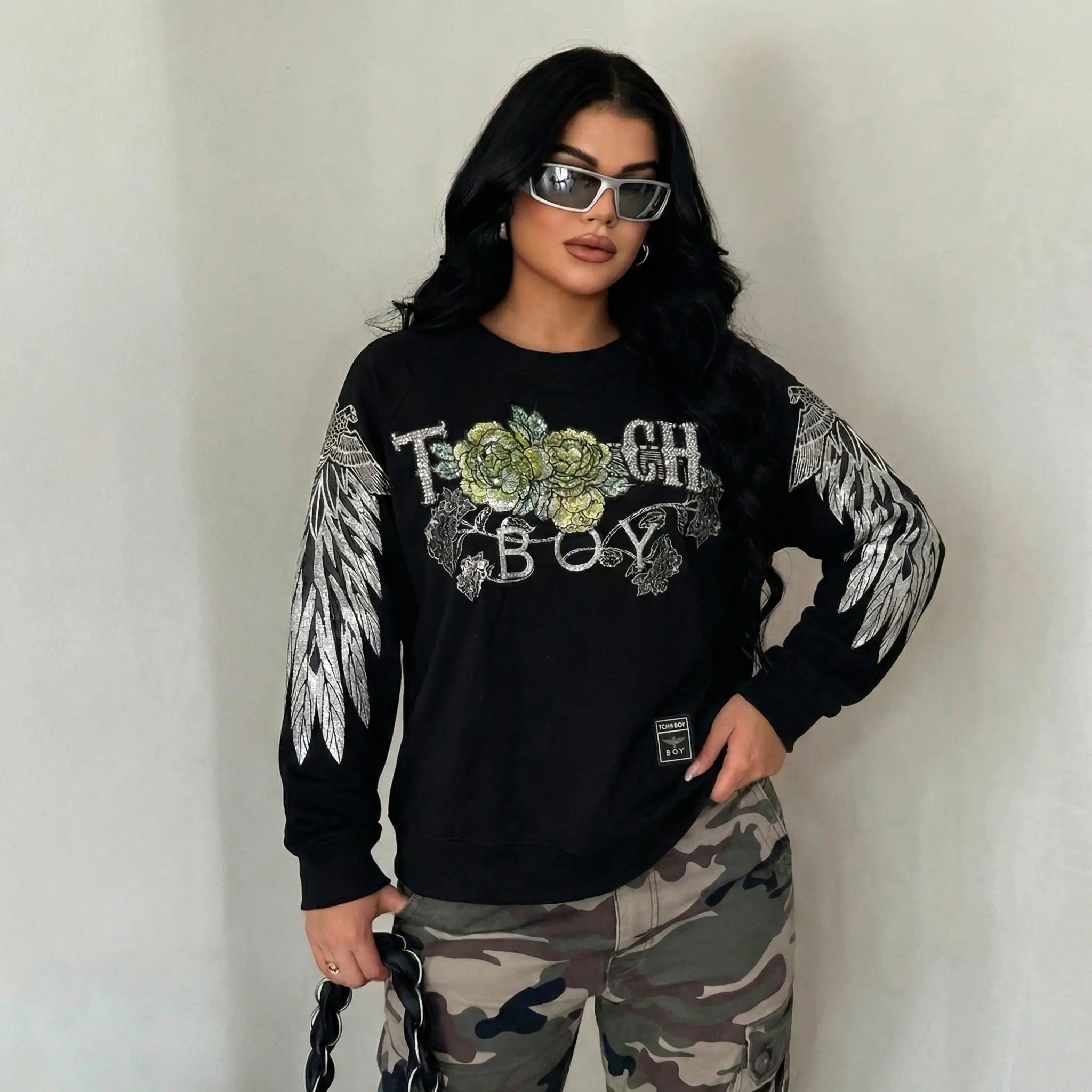 Person wearing a black sweatshirt with floral design and text, camouflage pants, and sunglasses on a plain background, TCH Rhinestone Peony Wing Graphic Crew Neck Sweatshirt