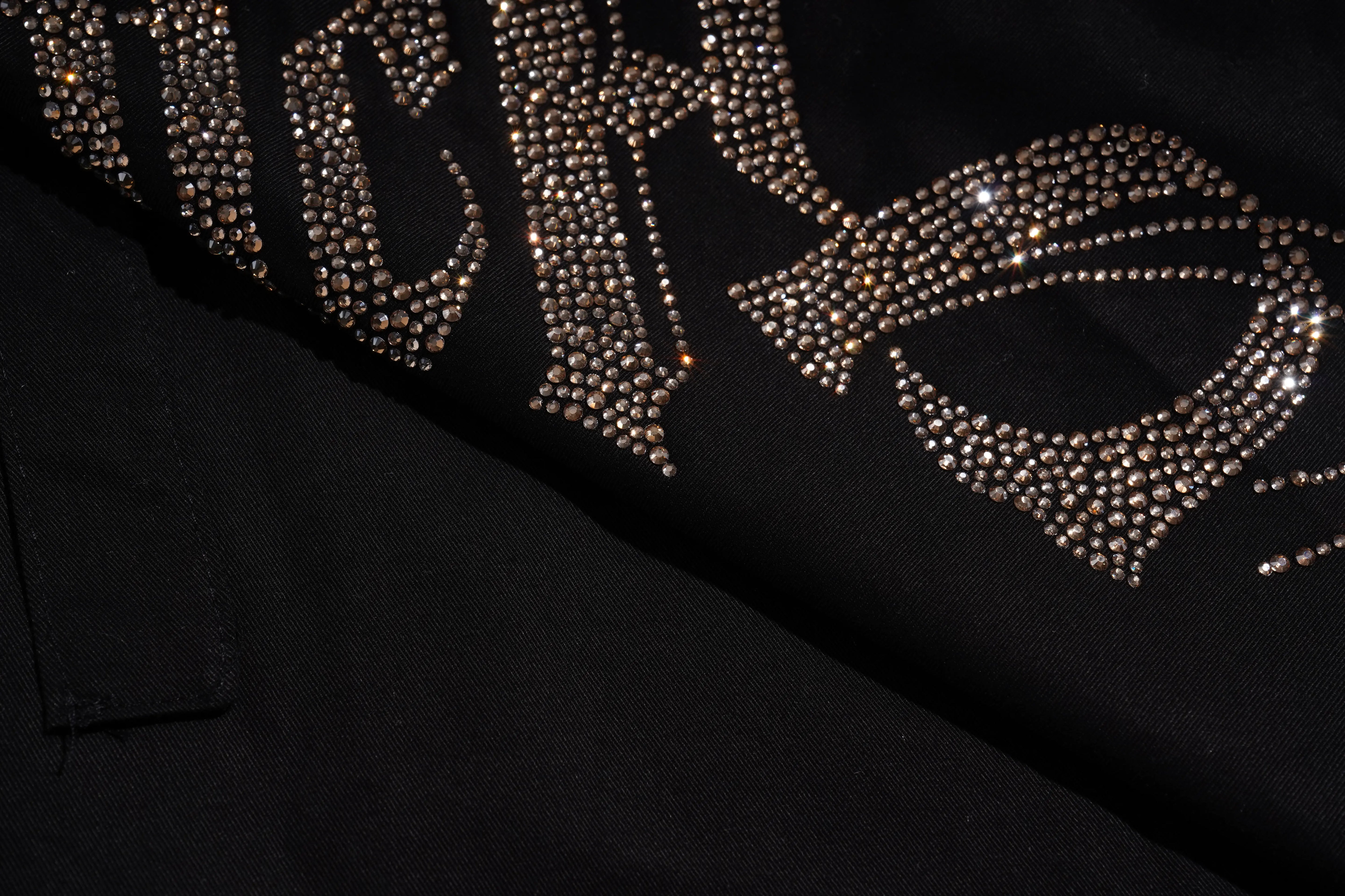 A close-up of a black garment featuring large, ornate lettering embellished with sparkling rhinestones in a champagne-gold tone. TCH Rhinestone Phoenix Cargo Wide-Leg Pants