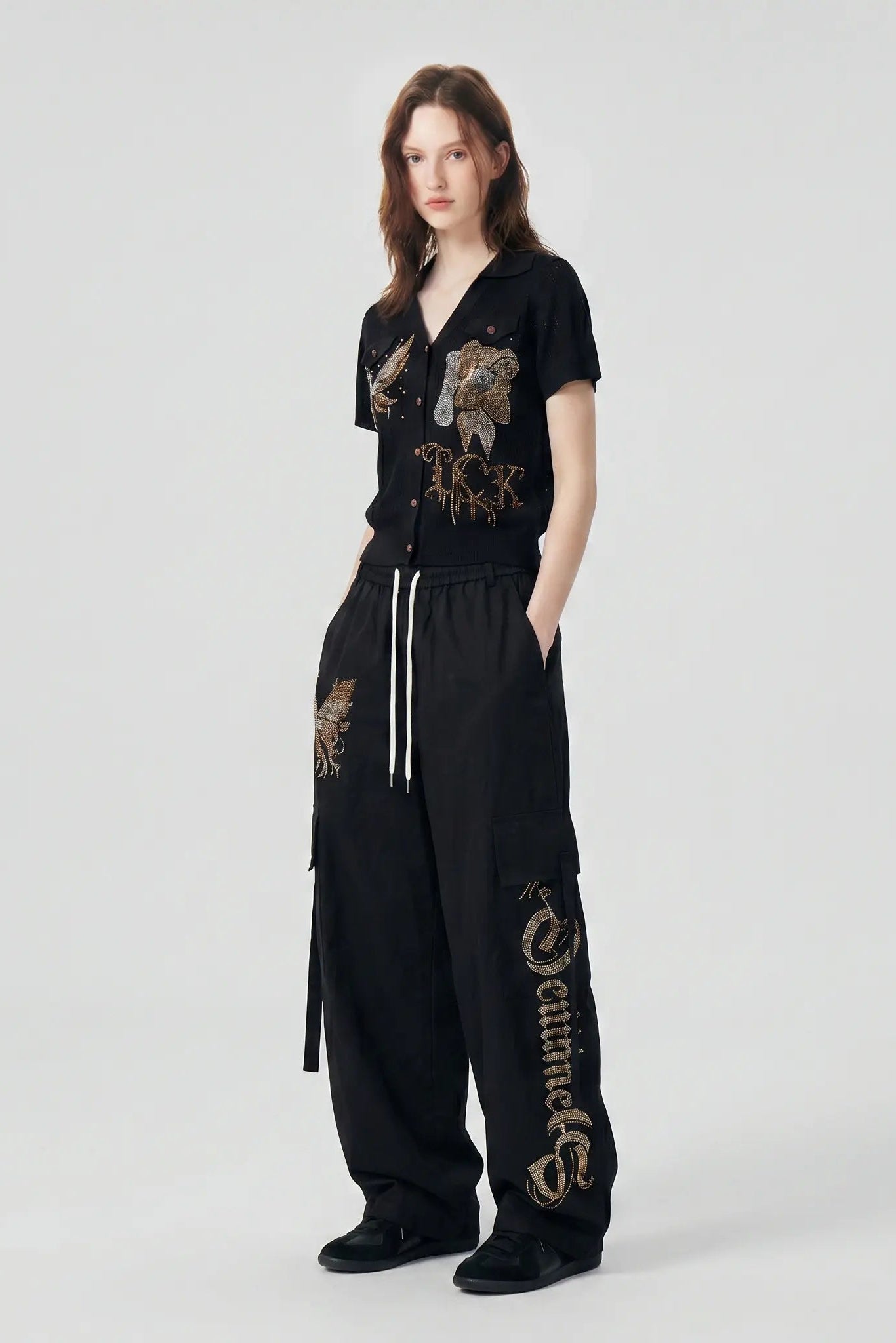 A model stands against a light gray studio background wearing a black short-sleeve button-front top and matching black wide-leg pants with embroidered details. The top has a lion graphic on the chest, and the pants have a vertical embroidered logo on one leg and a smaller matching graphic on the thigh. TCH Rhinestone Phoenix Cargo Wide-Leg Pants
