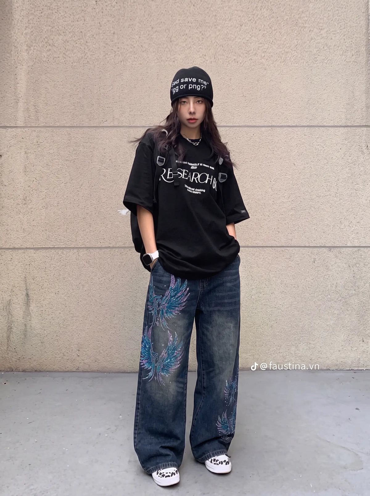 A person stands against a beige concrete wall wearing an oversized black graphic T-shirt, loose dark blue jeans with blue feather-like print details, a black beanie, and white sneakers. TCH Rhinestone Phoenix Wide-Leg Jeans