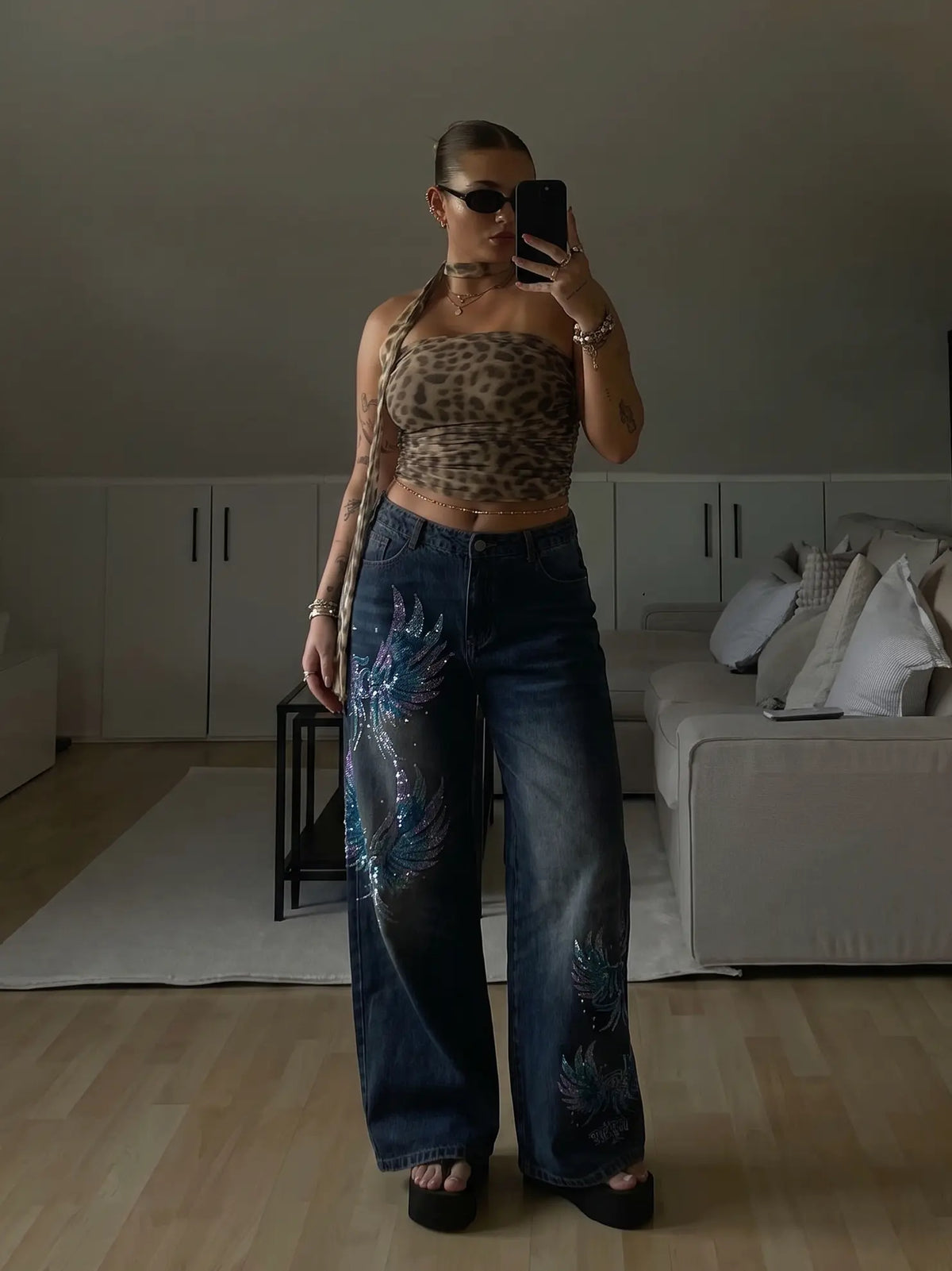 A woman takes a mirror selfie in a room, wearing a leopard-print halter crop top and dark blue wide-leg jeans with blue dragon embroidery on the legs, paired with black platform sandals. TCH Rhinestone Phoenix Wide-Leg Jeans