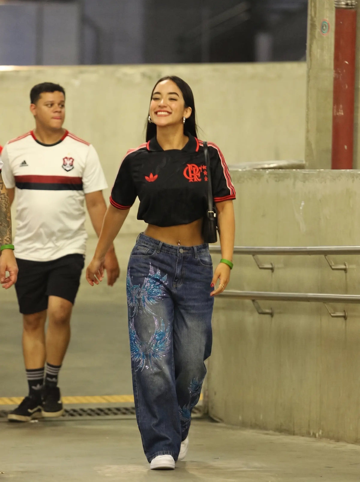 A woman walking in an indoor corridor wearing a black cropped Adidas Flamengo jersey top and blue jeans with a blue dragon graphic print. A man in a white Flamengo jersey walks behind her. TCH Rhinestone Phoenix Wide-Leg Jeans