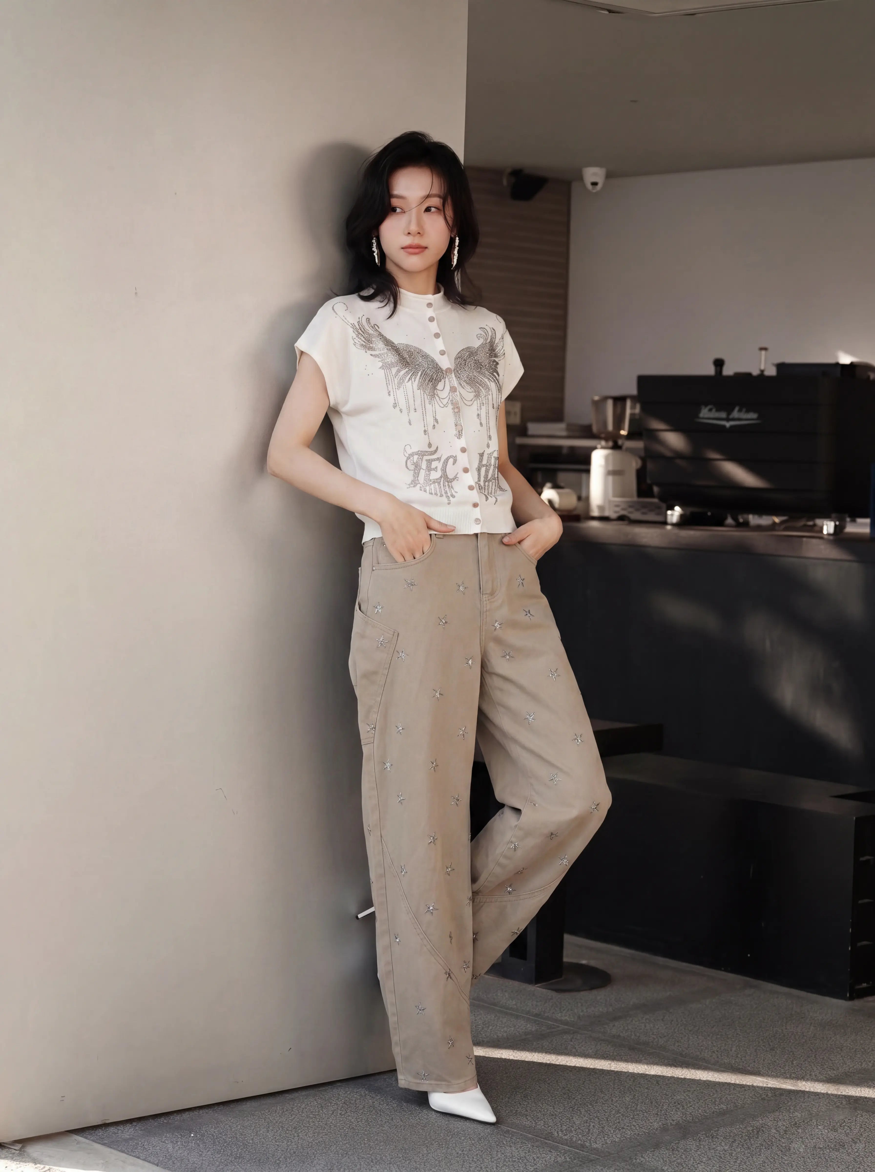 A woman stands in a modern indoor setting, leaning against a light gray wall. She is wearing a white short-sleeve t-shirt with a gray graphic print and beige wide-leg trousers with subtle silver embellishments. TCH Rhinestone Phoenix Wings Button Knit Top