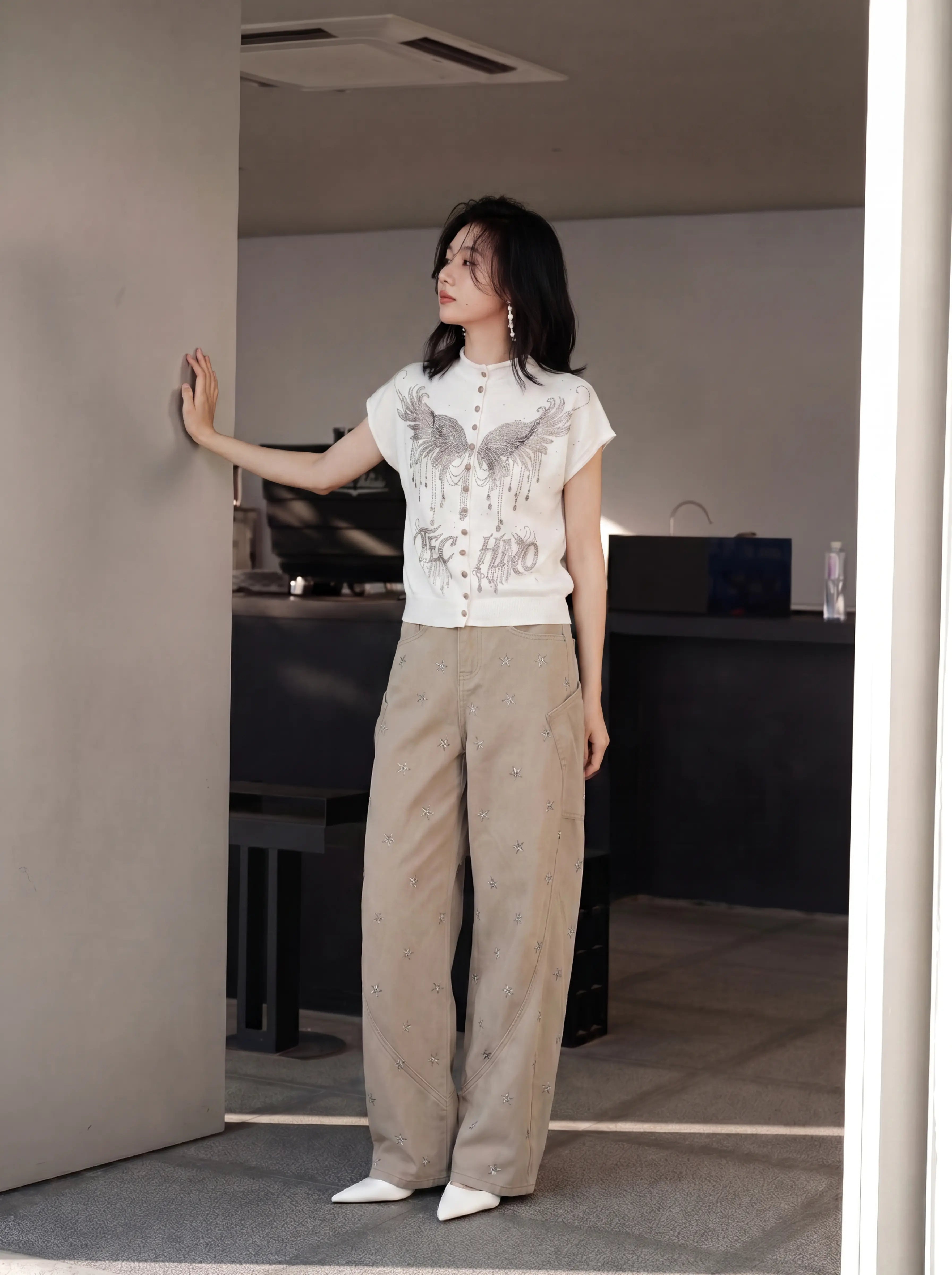 A woman stands in a doorway wearing a white short-sleeve button-front top with a large gray wing graphic and beige wide-leg pants, photographed in a modern indoor setting. TCH Rhinestone Phoenix Wings Button Knit Top