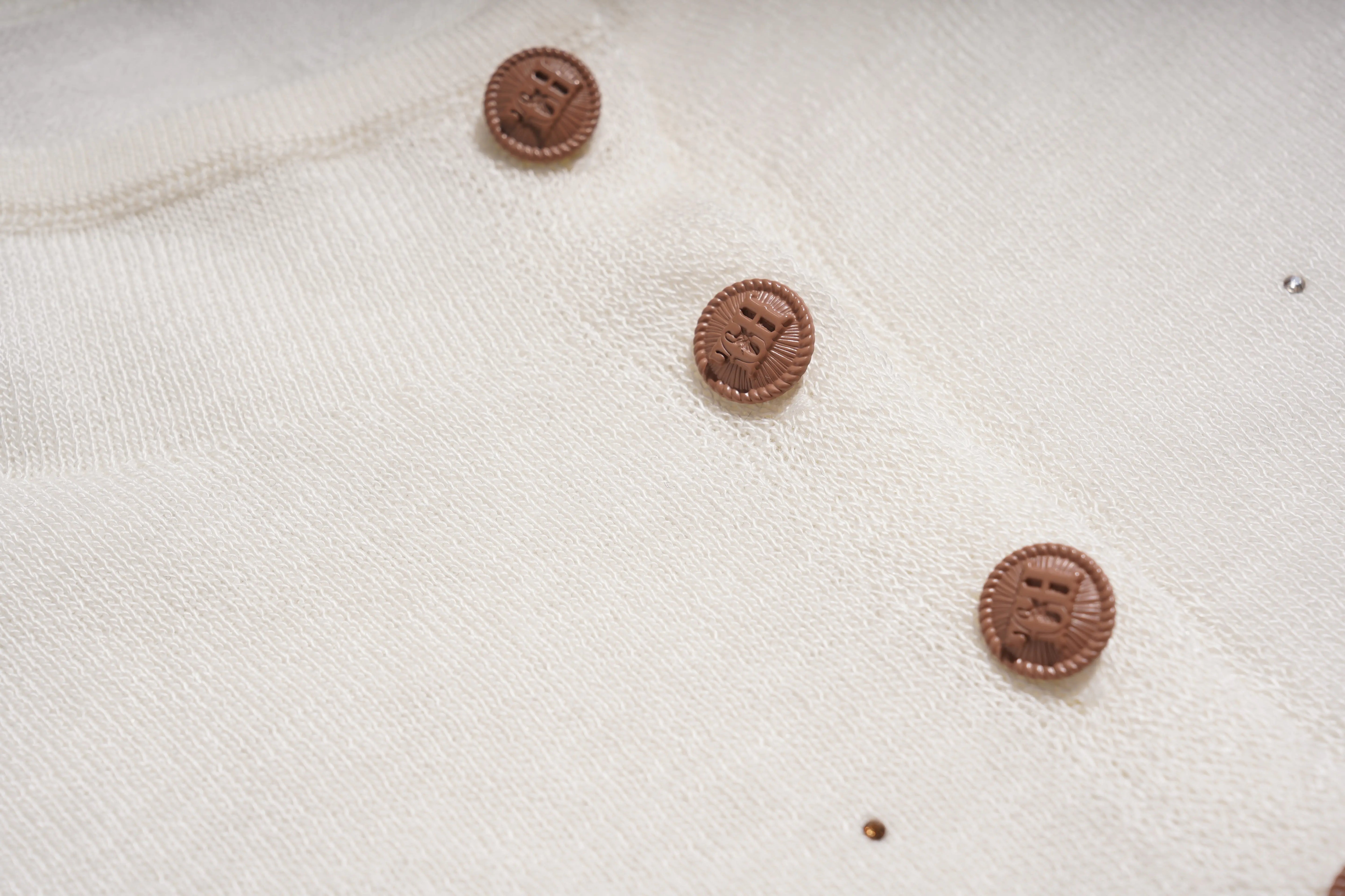Close-up of a cream-colored knit garment featuring three round, metallic bronze buttons with embossed lettering. TCH Rhinestone Phoenix Wings Button Knit Top