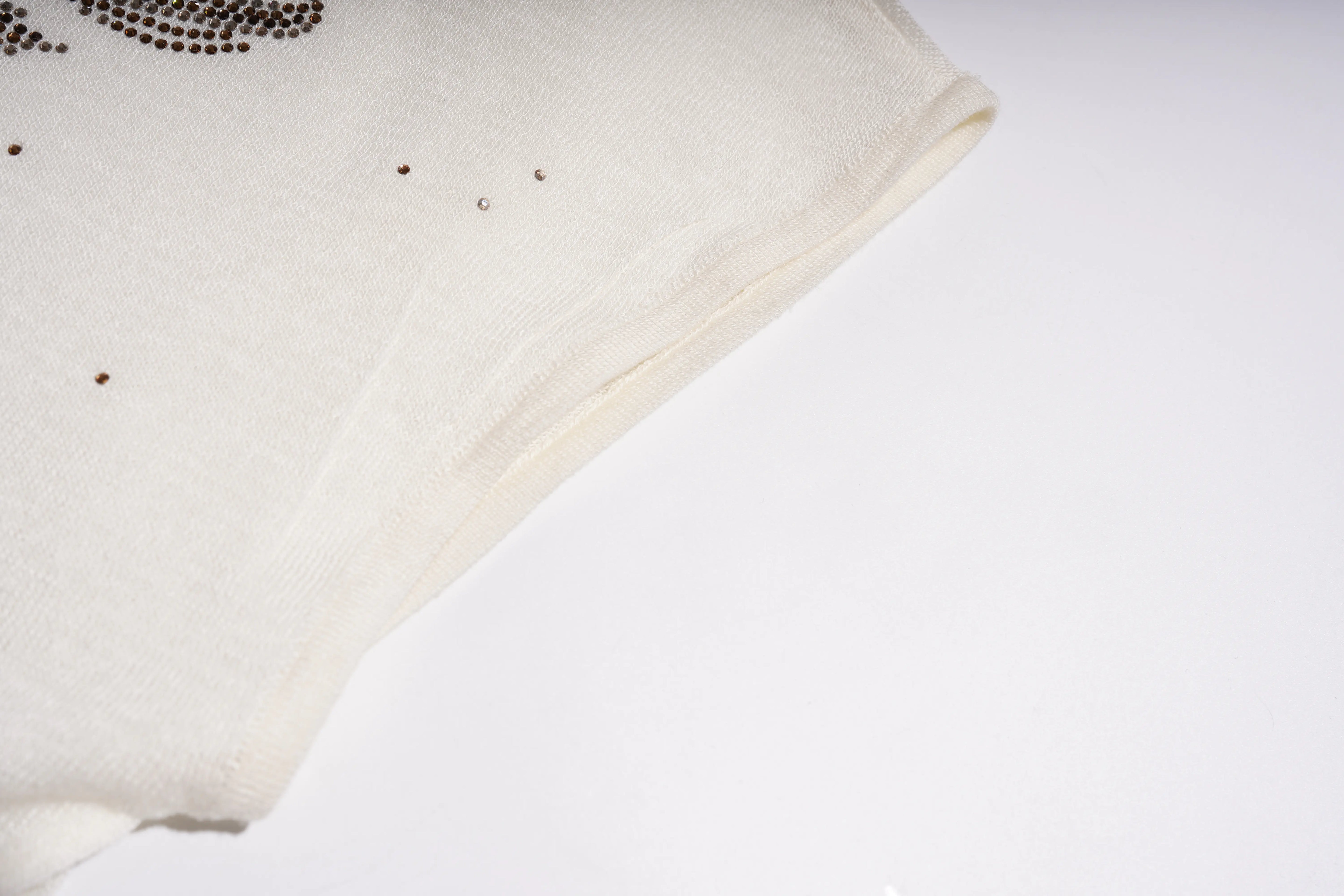 A close-up of a cream-colored knit fabric, likely a sweater or scarf, featuring small rhinestone embellishments on the left side, set against a plain white background. TCH Rhinestone Phoenix Wings Button Knit Top