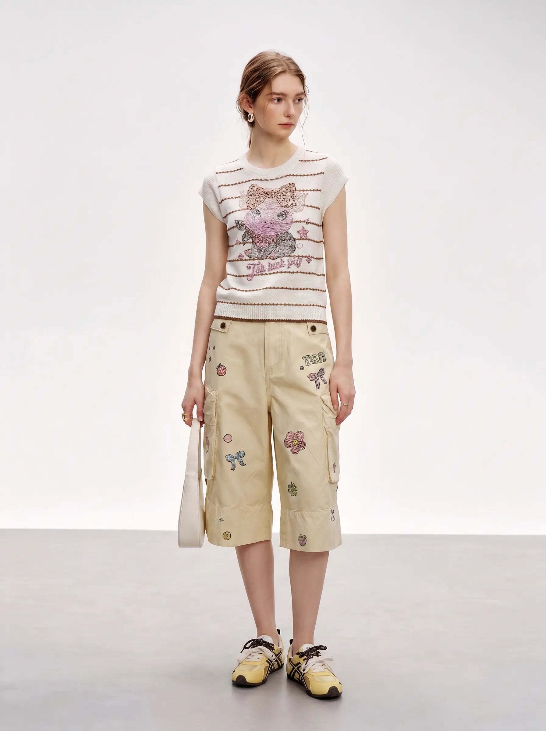 A model stands in a studio wearing a cream sleeveless knit top with a colorful graphic print and beige cargo shorts decorated with small floral patches, paired with yellow sneakers. TCH Rhinestone Piggy Bow Striped Knit Top