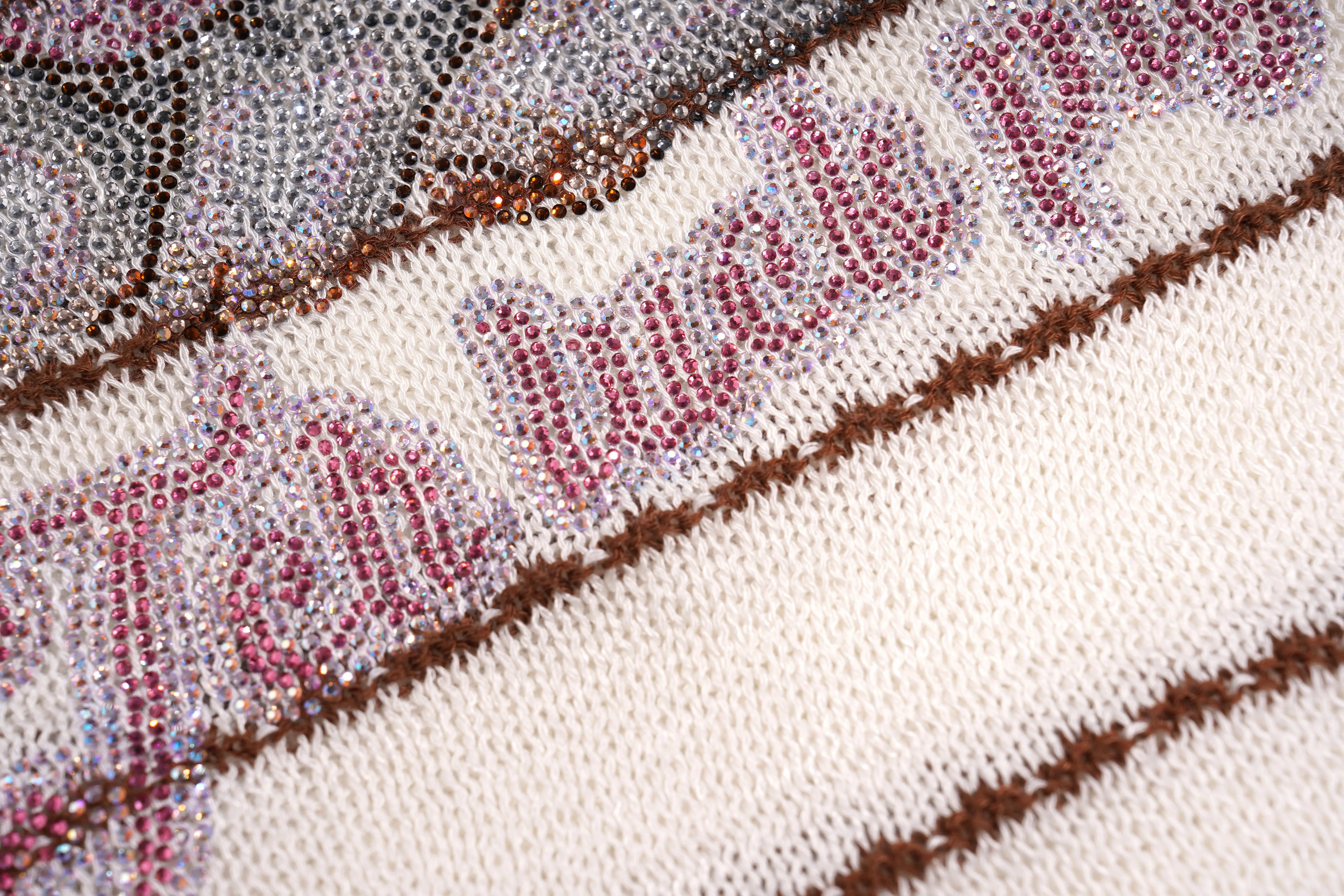 Close-up of a white knit fabric with pink embroidered gothic-style lettering outlined in brown, alongside sparkly silver and gold textured stripes. TCH Rhinestone Piggy Bow Striped Knit Top