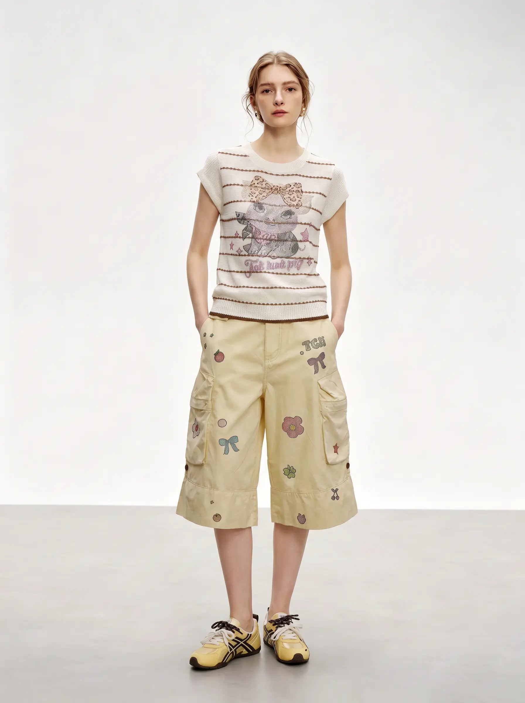 A model stands against a light gray studio background wearing a cream sleeveless knit top with horizontal stripes and a teddy bear graphic, paired with beige cargo shorts featuring colorful embroidered motifs, and yellow sneakers. TCH Rhinestone Piggy Bow Striped Knit Top