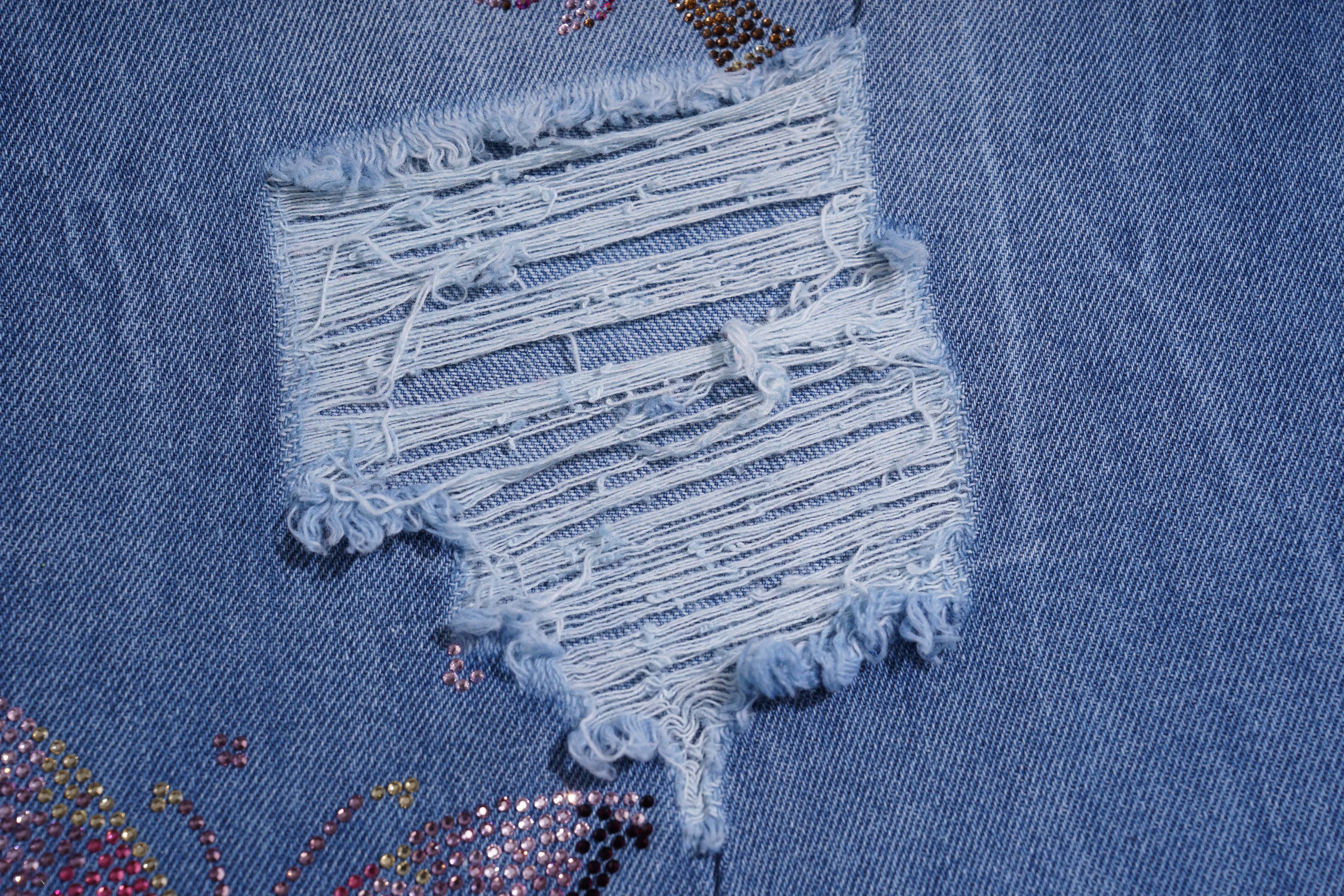 Close-up of a blue denim fabric with a frayed edge and decorative elements. TCH Rhinestone Plum Blossom Tree Distressed Denim Jeans
