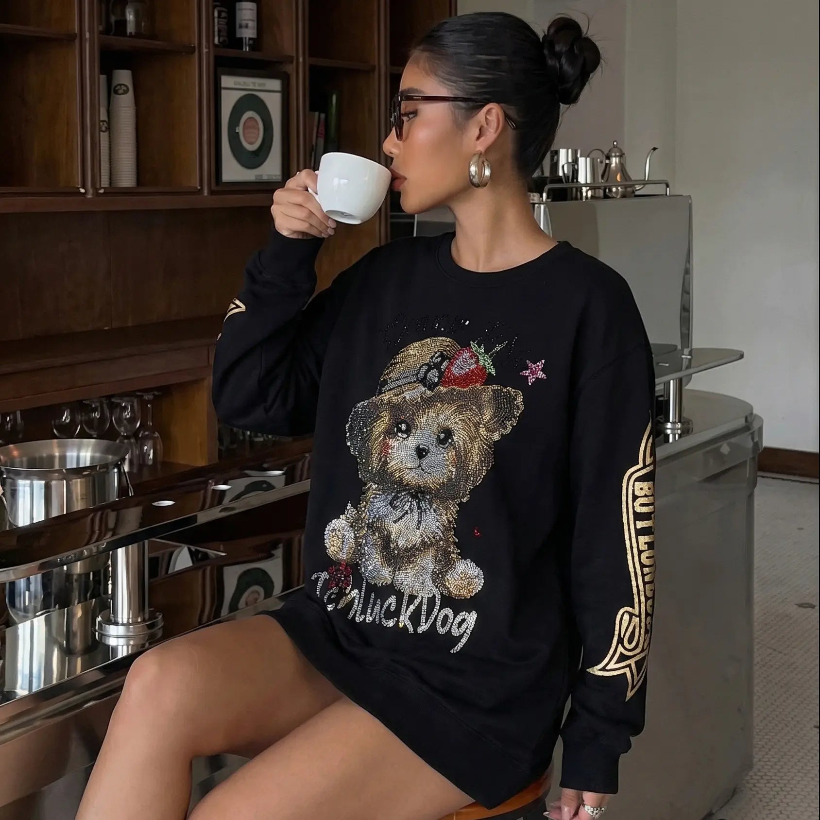Woman wearing a black sweatshirt with a teddy bear design, sitting in a kitchen. TCH Rhinestone Puppy Graphic Sweatshirt-1