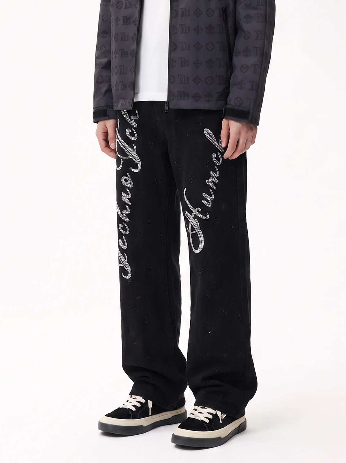 A person wearing black wide-leg Techno Funk pants with large white script logos down both legs, styled with a dark patterned jacket, white shirt, and black sneakers with white soles, against a plain white studio background. TCH Rhinestone Script Graphic Wide-Leg Jeans