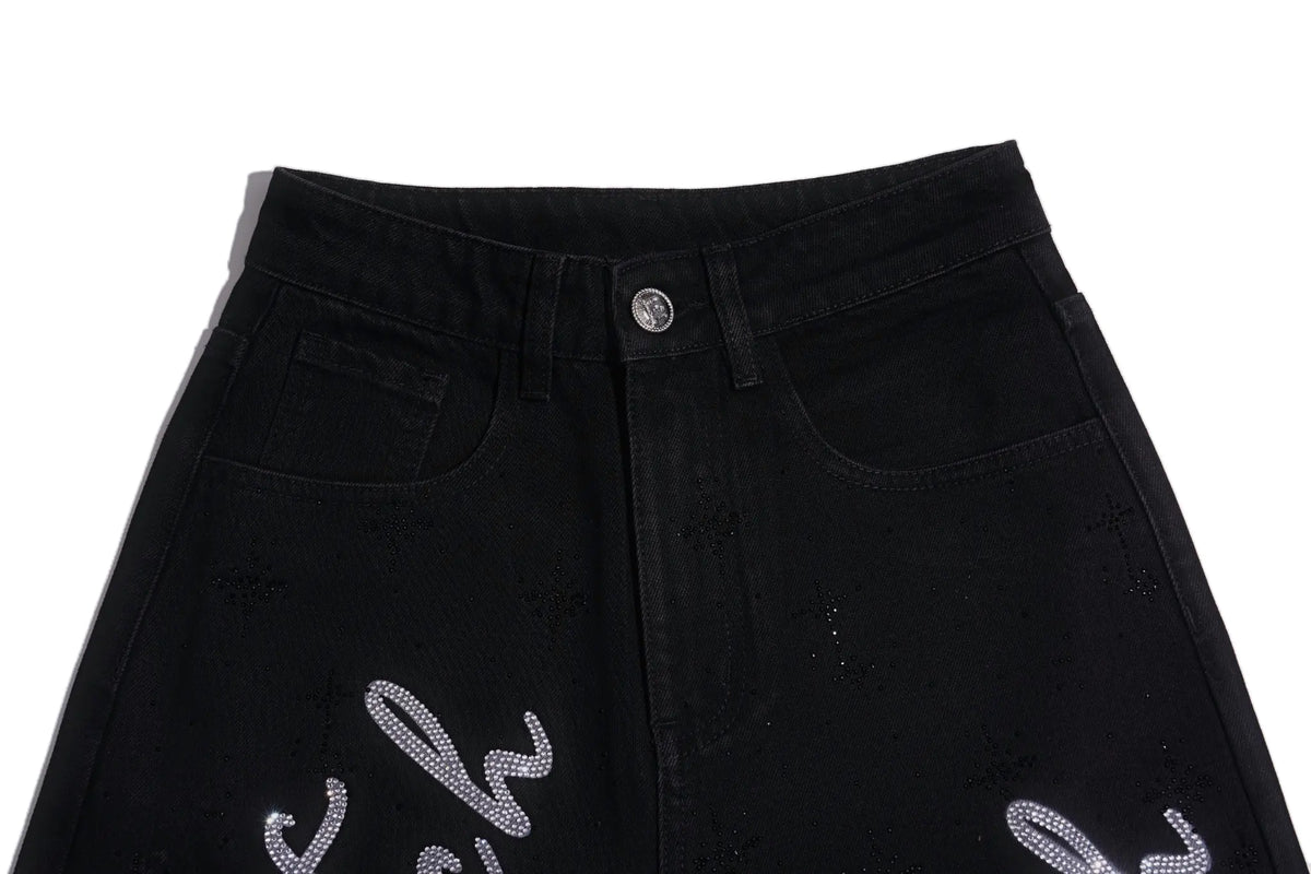 Close-up of black denim jeans showing the front waistband, button, belt loops, and decorative silver rhinestone embroidery on the lower legs, set against a plain white background. TCH Rhinestone Script Graphic Wide-Leg Jeans