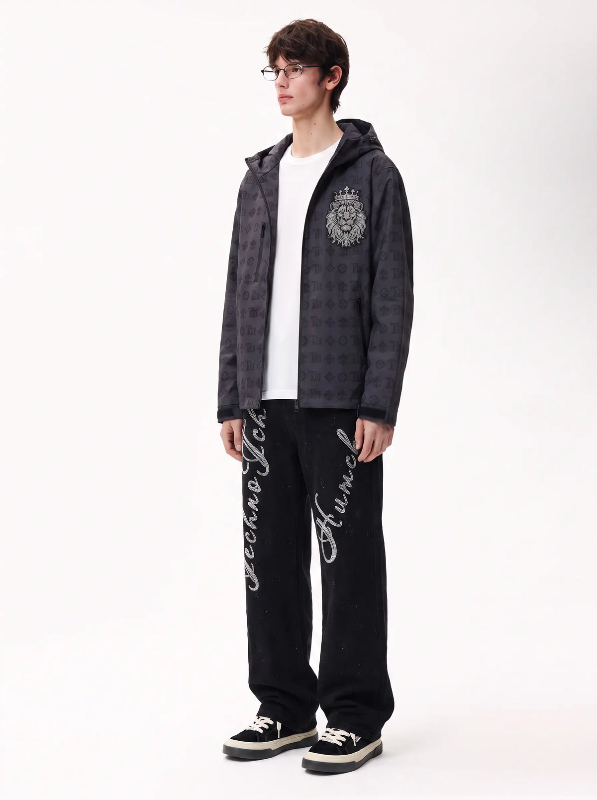 A model stands against a white studio background wearing a dark patterned zip-up hoodie with a lion crest on the chest, a white t-shirt, black sweatpants with white script graphics, and black sneakers. TCH Rhinestone Script Graphic Wide-Leg Jeans