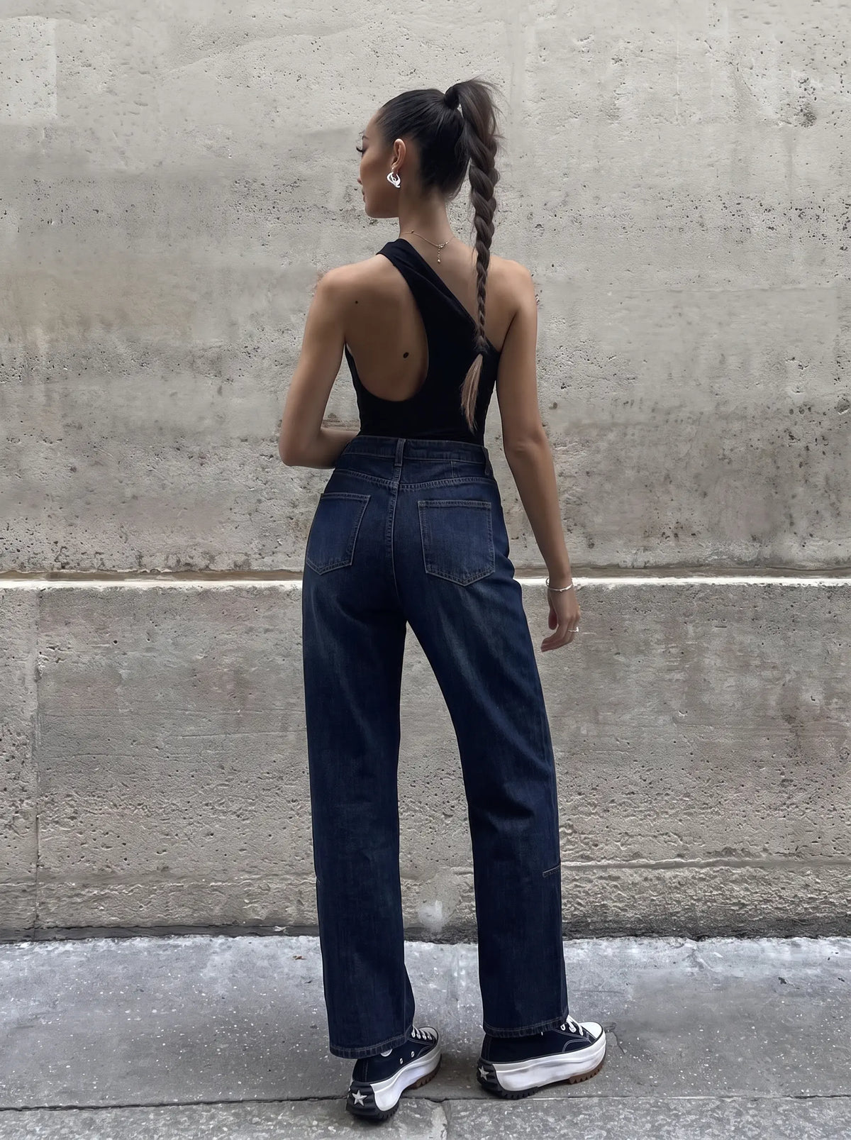 A woman stands with her back to the camera wearing a black one-shoulder backless top, dark blue wide-leg jeans, and black platform sneakers, posing against a textured concrete wall. TCH Rhinestone Shooting Star Galaxy Jeans