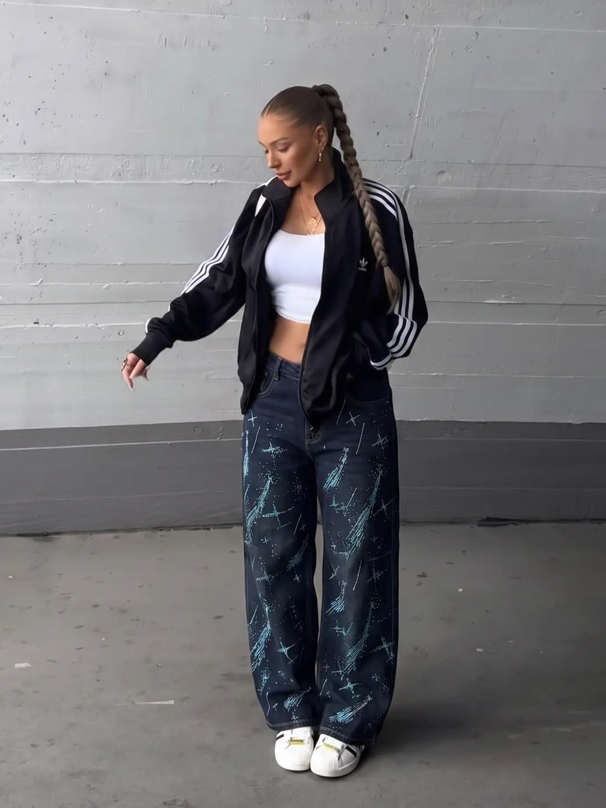 A woman stands in a studio wearing a black Adidas track jacket with white stripes, a white crop top, dark blue wide-leg jeans with a blue marble pattern, and white sneakers. TCH Rhinestone Shooting Star Galaxy Jeans