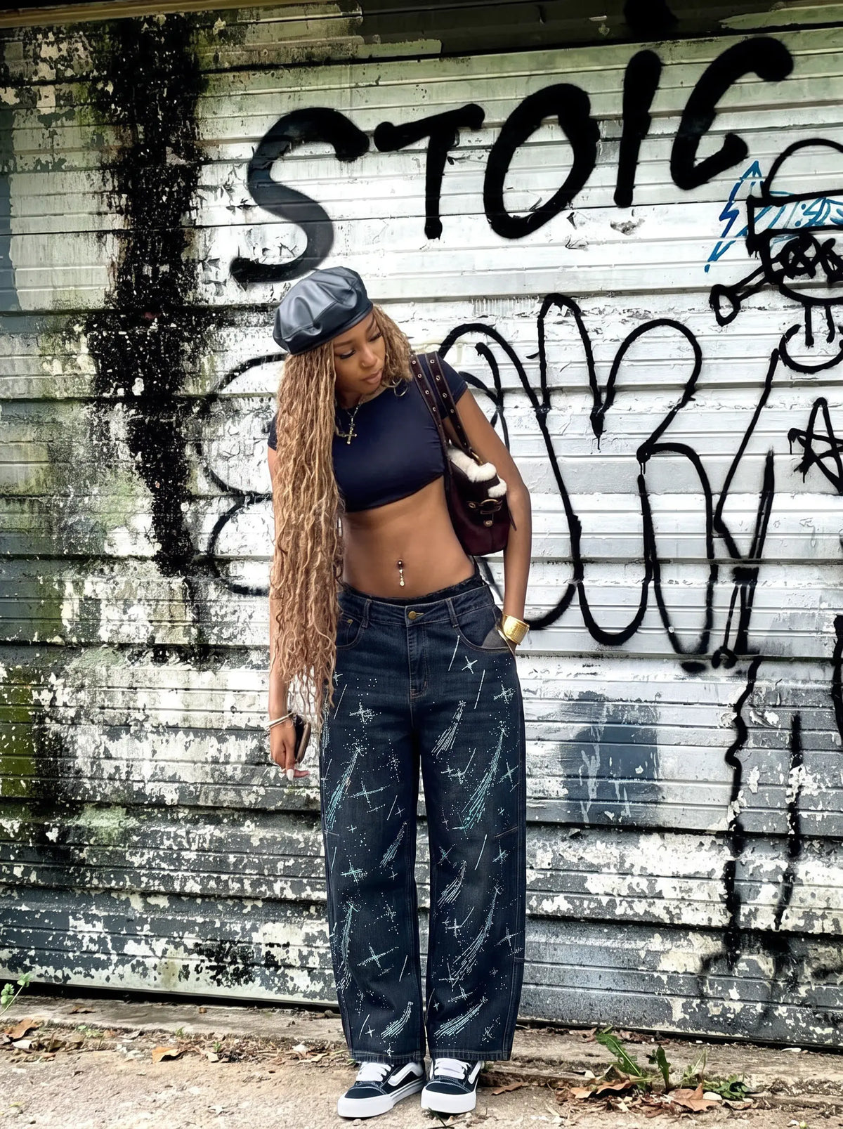 A woman stands in front of a graffiti-covered metal wall, wearing a dark navy crop top, wide-leg dark blue jeans with white paint splatters, a black beret, and white sneakers. TCH Rhinestone Shooting Star Galaxy Jeans