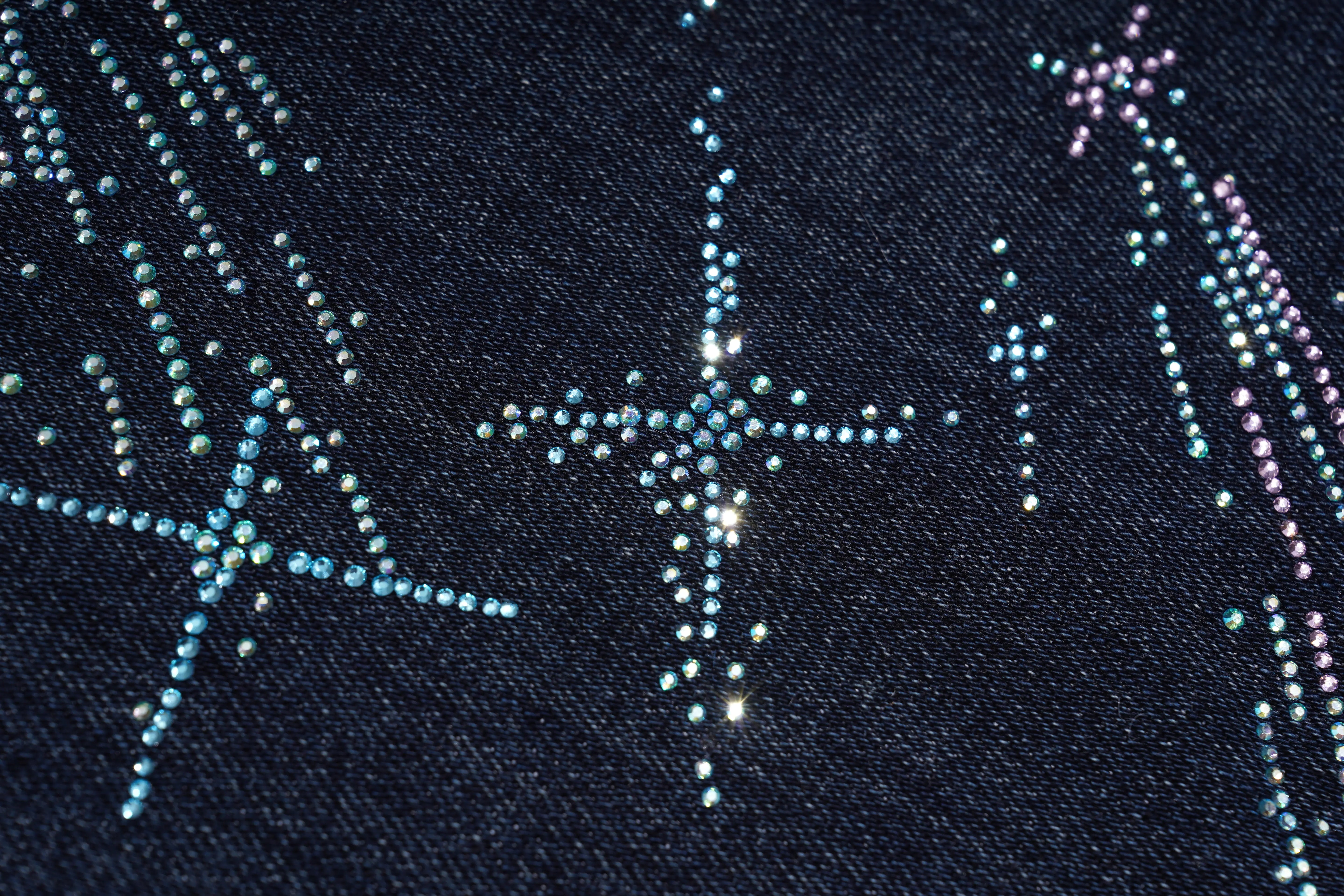 Close-up of a dark navy fabric with decorative rhinestone star and shooting star patterns in light blue, silver, and pink. TCH Rhinestone Shooting Star Galaxy Jeans