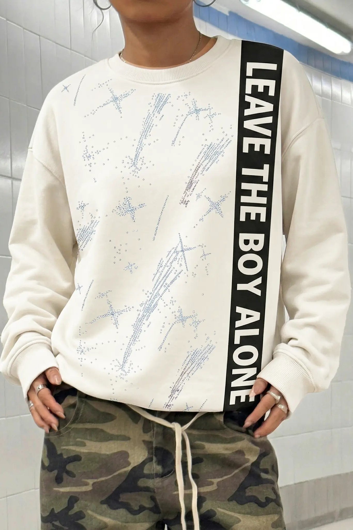 Person wearing a white sweatshirt with text and camouflage pants in a tiled room. TCH Rhinestone Shooting Star Sweatshirt