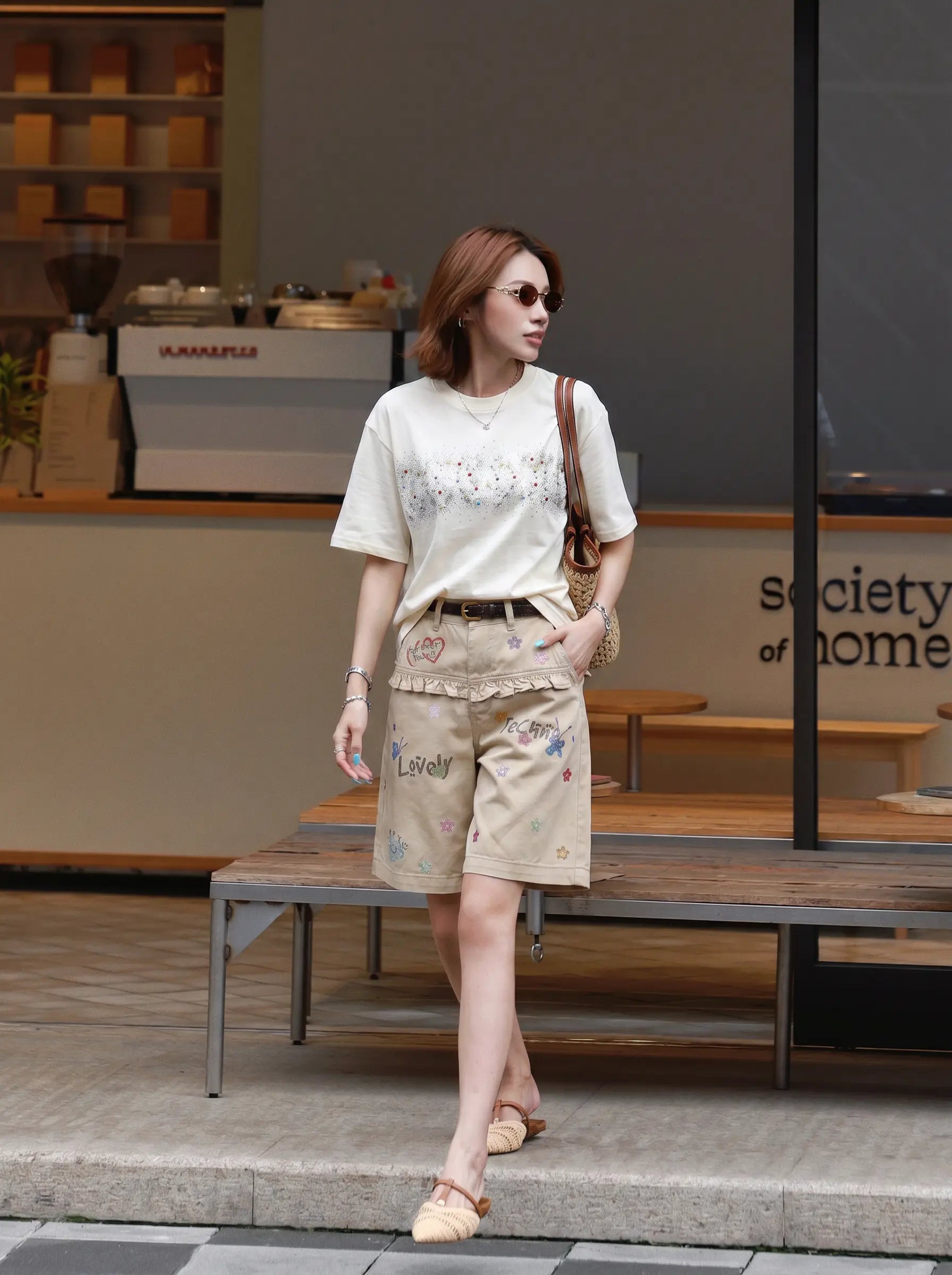 A woman wearing a cream graphic t-shirt, beige embroidered shorts, and beige flat sandals, walking in front of a cafe storefront. TCH Rhinestone Sparkle Crystal Graphic White T-Shirt