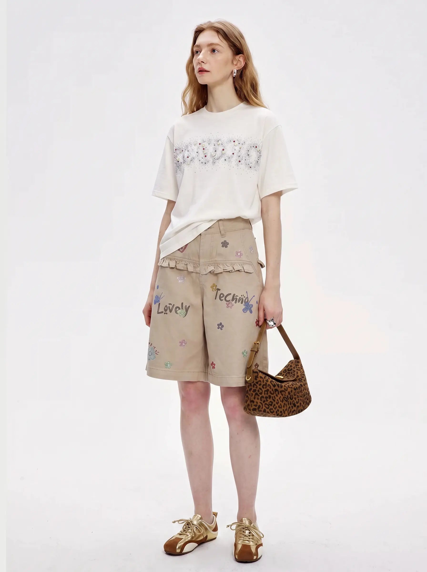 A model stands against a plain white studio background wearing a white graphic t-shirt, beige shorts with colorful text and floral prints, and holding a brown woven handbag. TCH Rhinestone Sparkle Crystal Graphic White T-Shirt