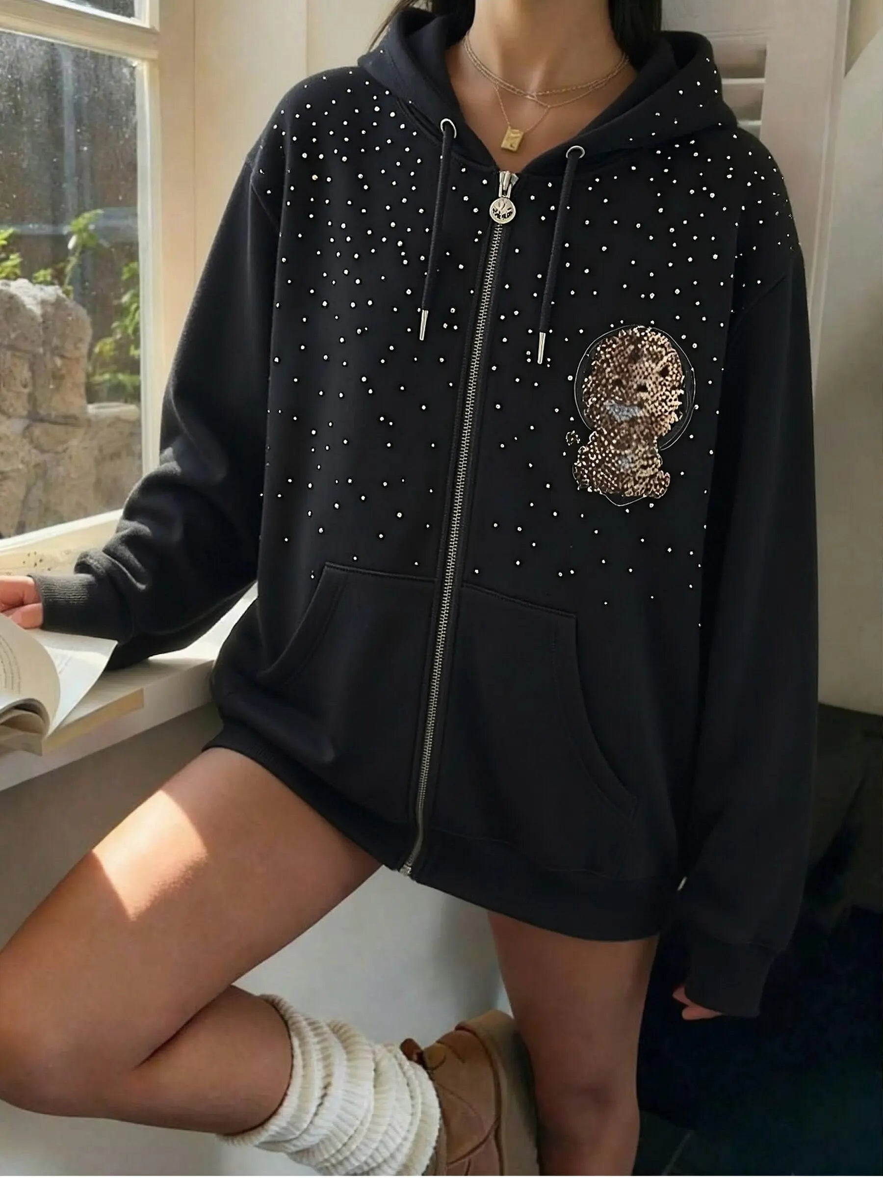 Person wearing a black hoodie with a lion emblem and rhinestone details, sitting by a window. TCH Rhinestone Starry Sky Lion Hoodie
