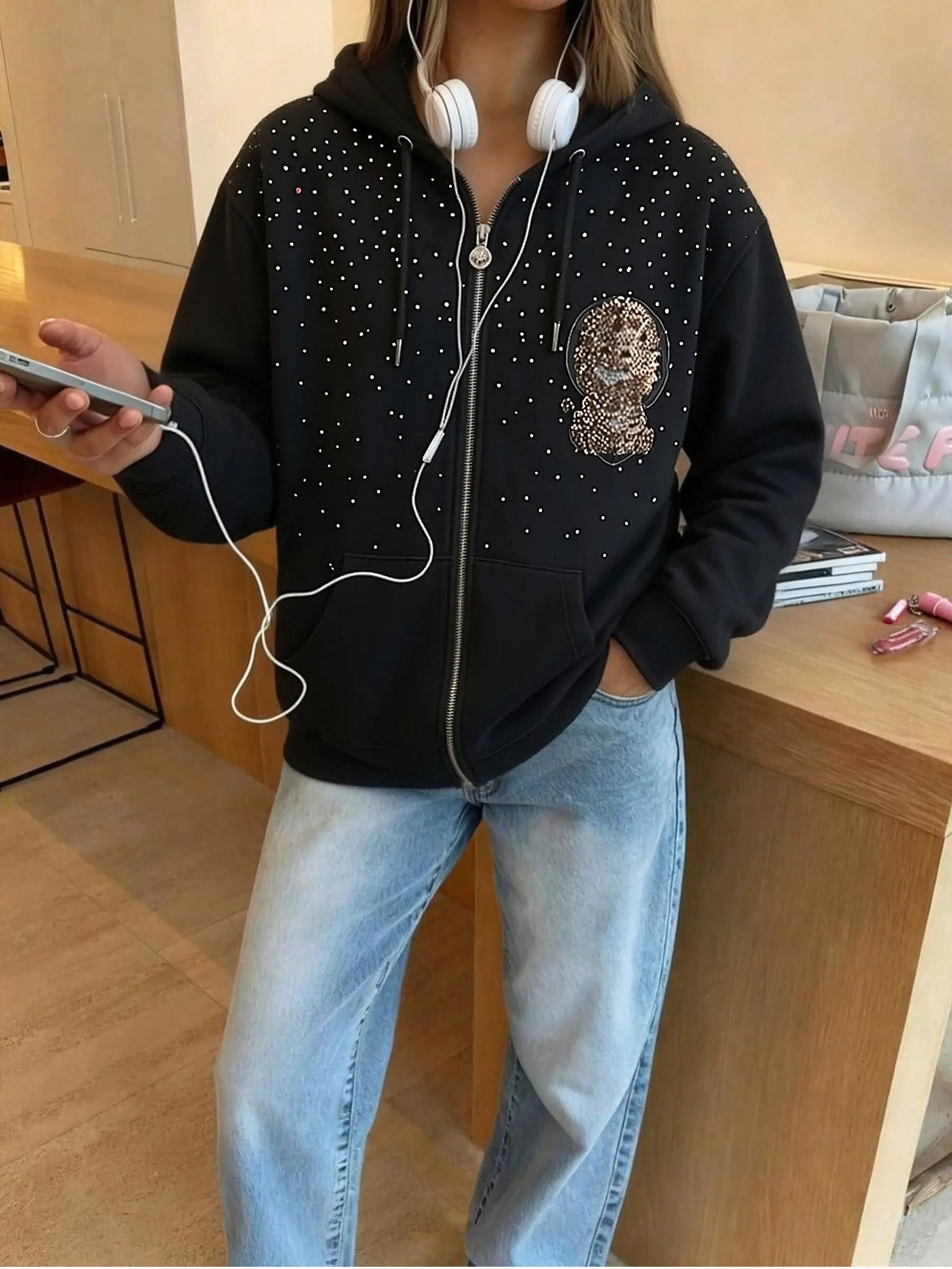 Person wearing a black hoodie with a face design and blue jeans, holding a phone and earphones indoors. TCH Rhinestone Starry Sky Lion Hoodie