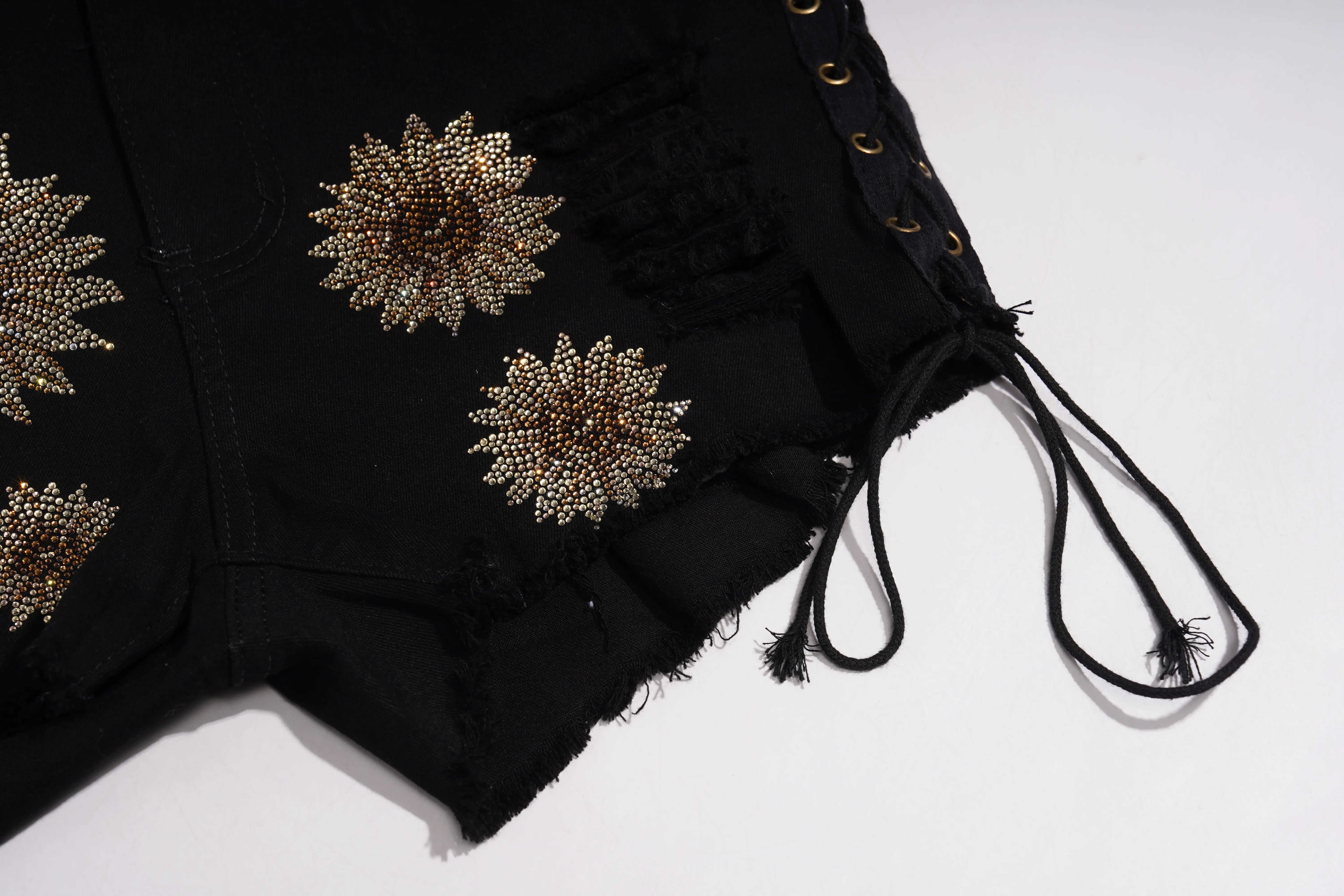 Close-up of black shorts with gold beaded starburst embellishments and side lacing with drawstrings, laid flat on a white background. TCH Rhinestone Sunflower Distressed Denim Shorts