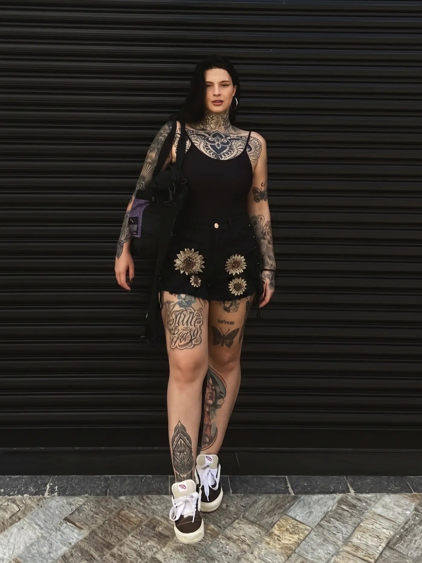 A tattooed woman stands in front of a black metal shutter wearing a black sleeveless mini dress with sunflower embroidery, white sneakers, and carrying a black shoulder bag. TCH Rhinestone Sunflower Distressed Denim Shorts