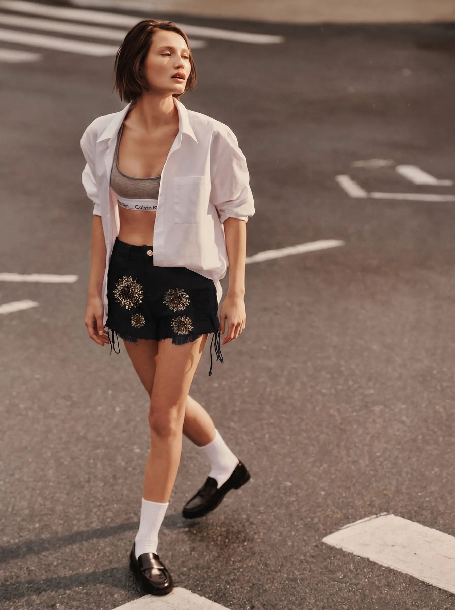 A woman walking on an asphalt street crosswalk wearing a white button-down shirt, a gray sports bra, black denim shorts with sunflower embroidery, white socks, and black loafers. TCH Rhinestone Sunflower Distressed Denim Shorts