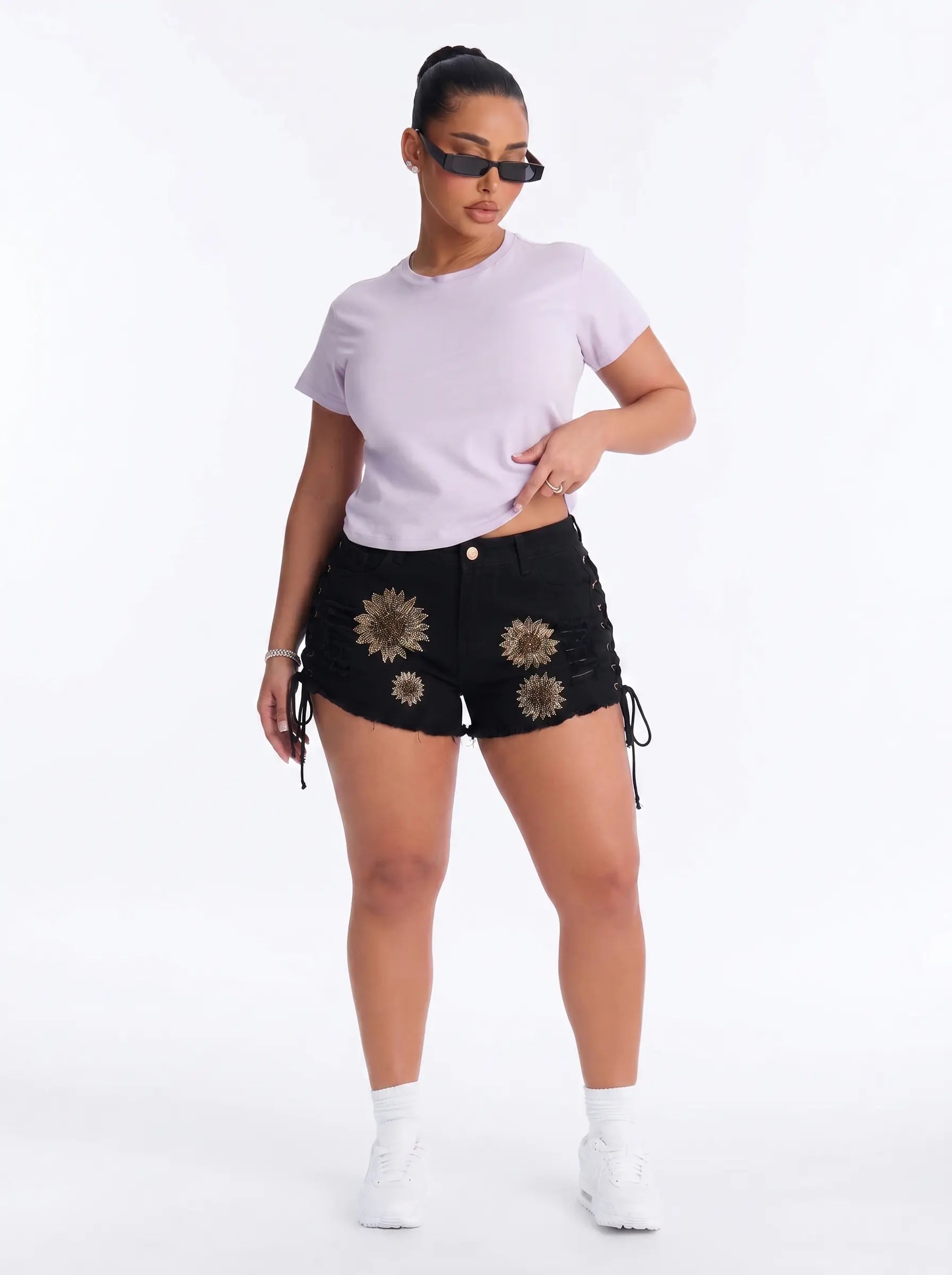 A woman modeling black denim shorts with gold floral embroidery and side laces, paired with a light purple cropped t-shirt, white sneakers, and sunglasses against a white studio background. TCH Rhinestone Sunflower Distressed Denim Shorts
