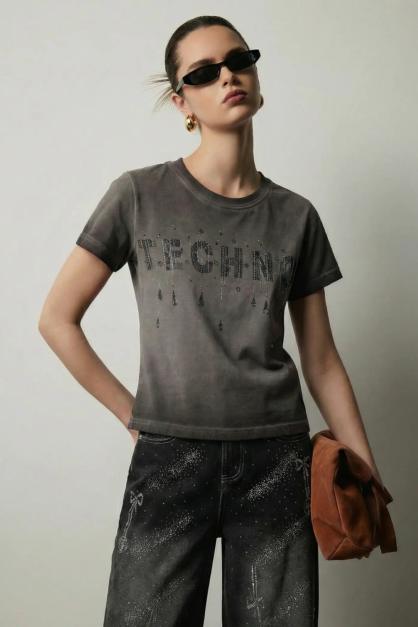 A model wearing a dark gray graphic t-shirt with distressed lettering, paired with dark gray pants and holding a brown clutch bag, set against a plain light gray background. TCH Rhinestone TECHNO Graphic Washed T-Shirt