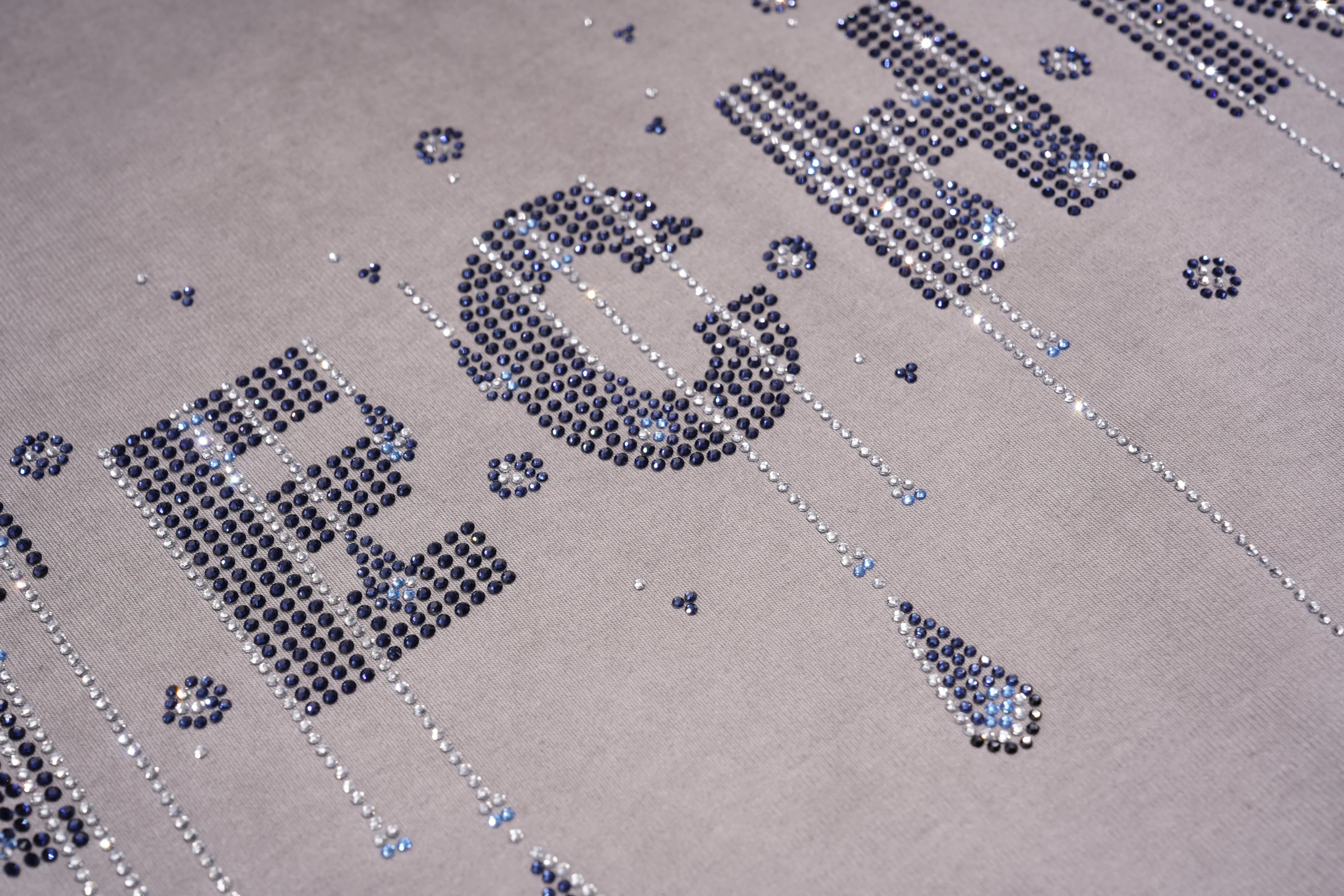 A close-up of a light gray fabric featuring large, sparkling rhinestone letters spelling 'RICH' in silver and blue tones, with decorative teardrop and dot embellishments. TCH Rhinestone TECHNO Graphic Washed T-Shirt