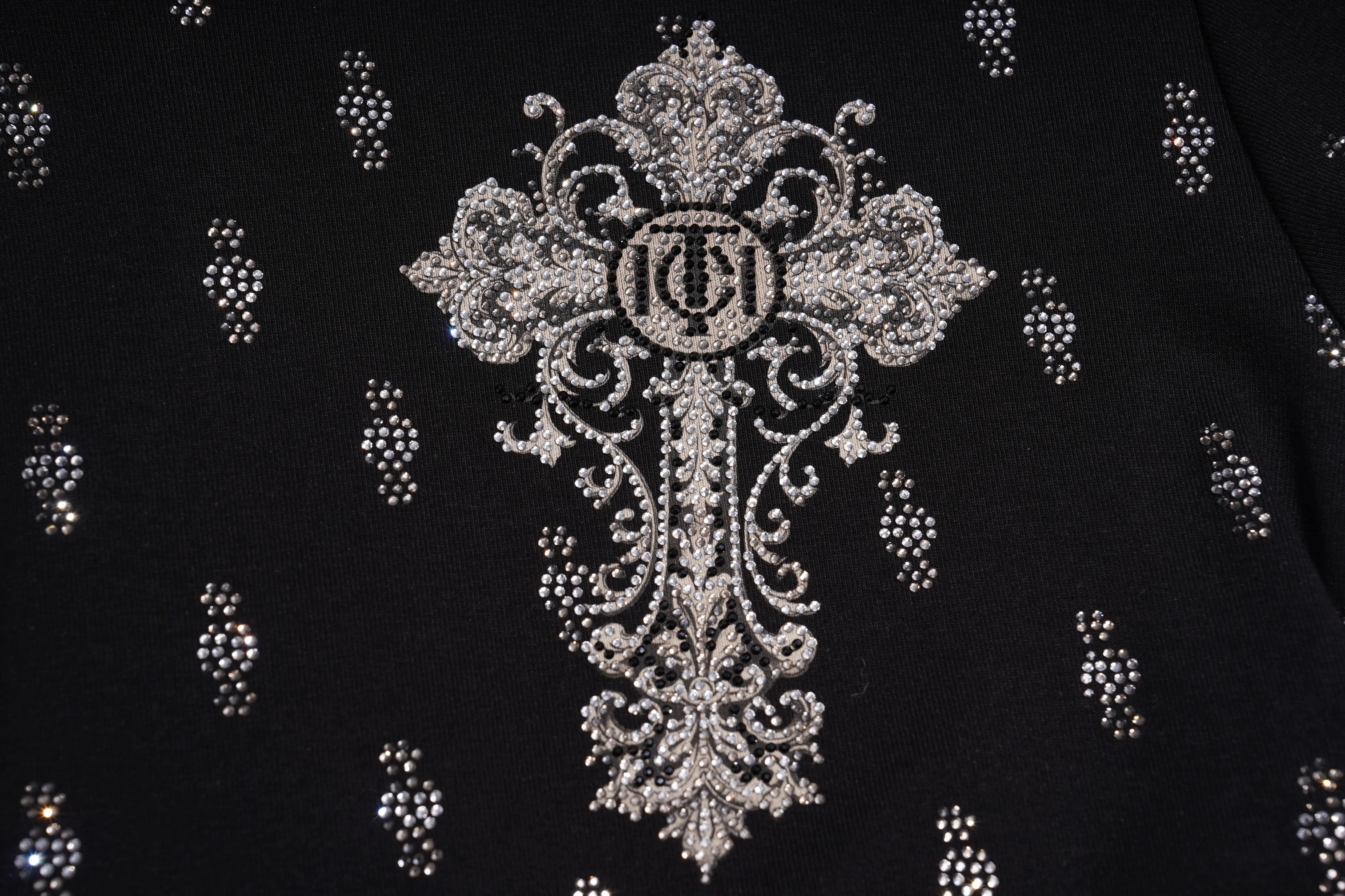 A close-up of a black garment featuring a large ornate silver rhinestone cross motif with a central monogram logo, surrounded by smaller matching silver embellishments. TCH Rhinestone Techno Cross Graphic Black T-Shirt