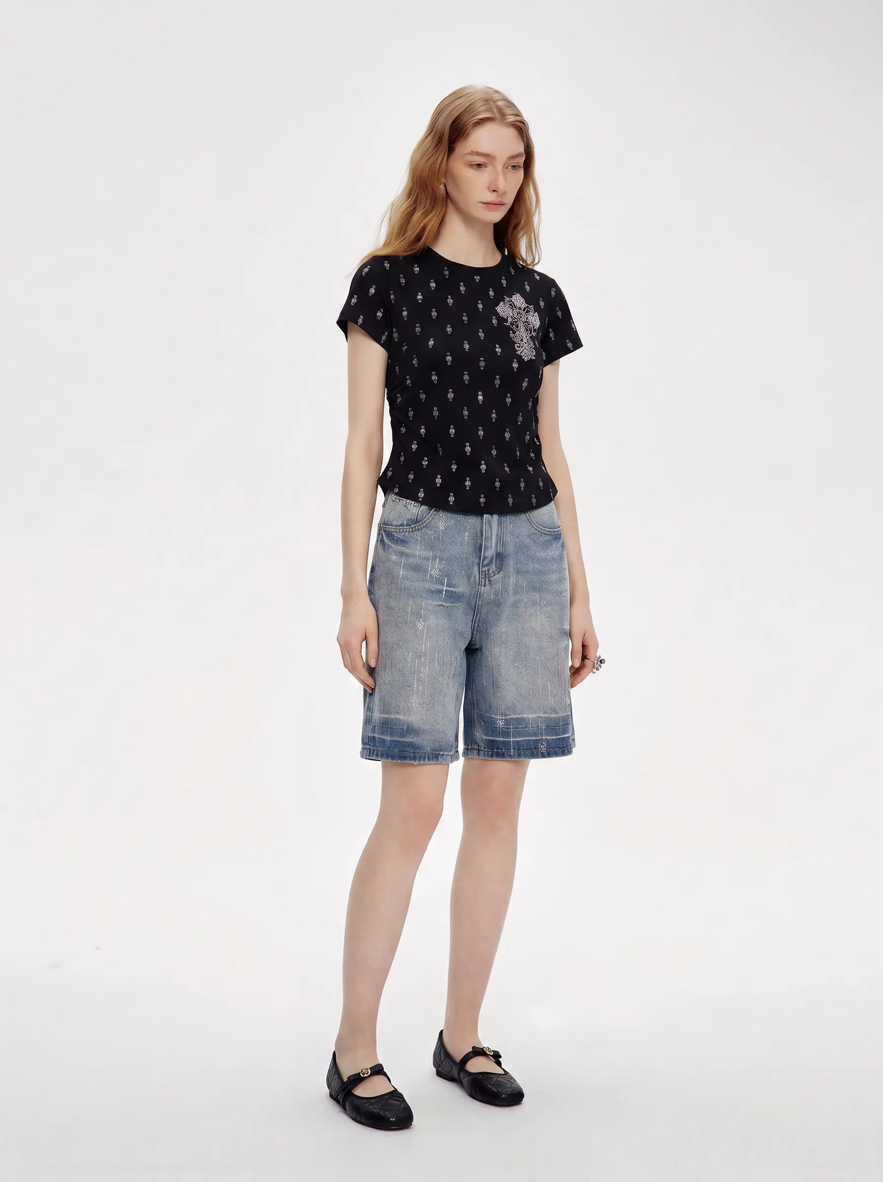 A woman modeling a black short-sleeve t-shirt with a small repeating pattern and a gray graphic on the left chest, paired with light-wash denim shorts and black Mary Jane shoes, against a plain white studio background. TCH Rhinestone Techno Cross Graphic Black T-Shirt